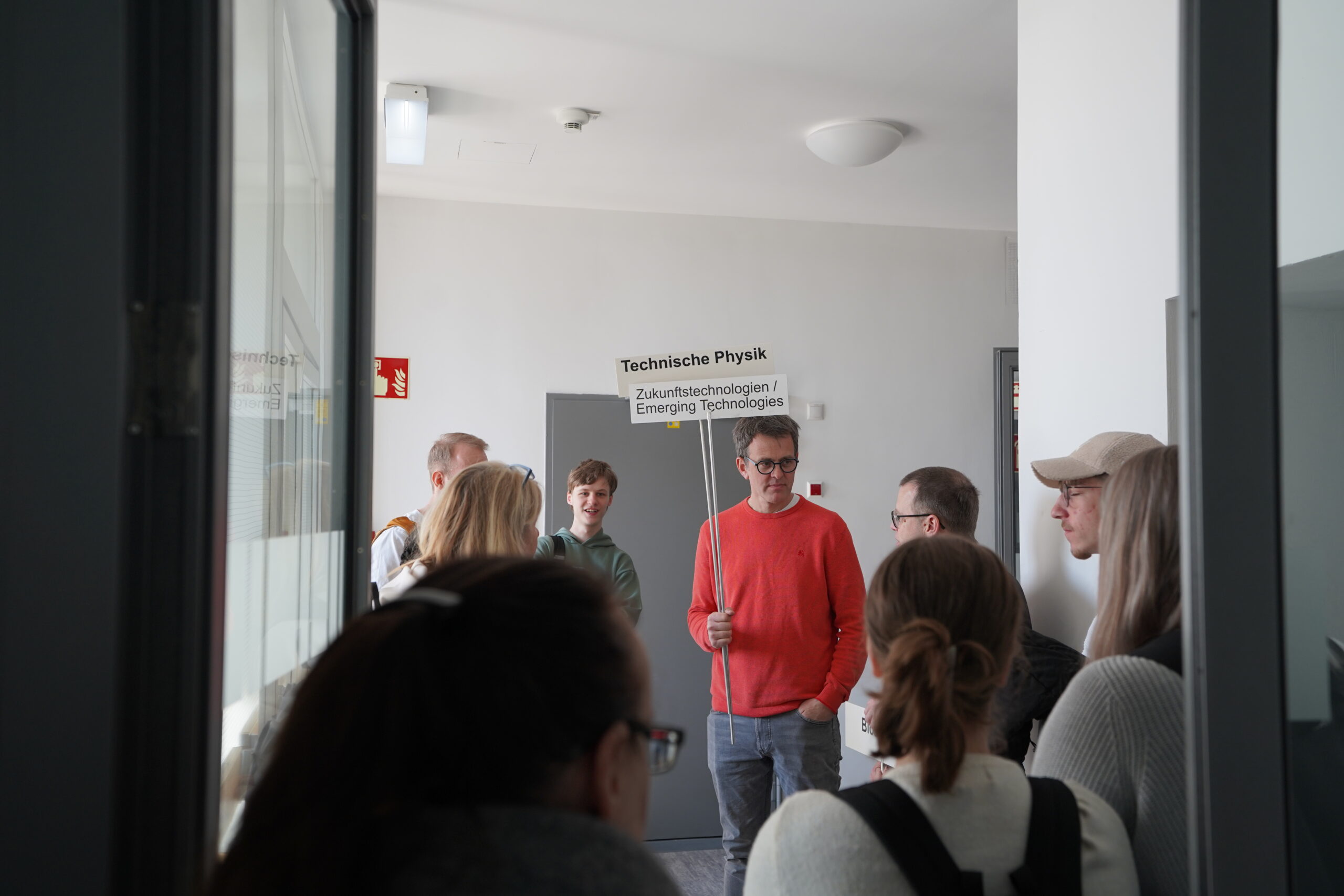 Eine Gruppe von Menschen steht in einem Flur des Hochschul-Campus und hört einem Mann in einem roten Pullover zu, der ein Schild mit der Aufschrift "Technische Physik / Zukunftstechnologien | Emerging Technologies" im Rahmen des Studieninfo-Programms hält. at Hochschule Coburg