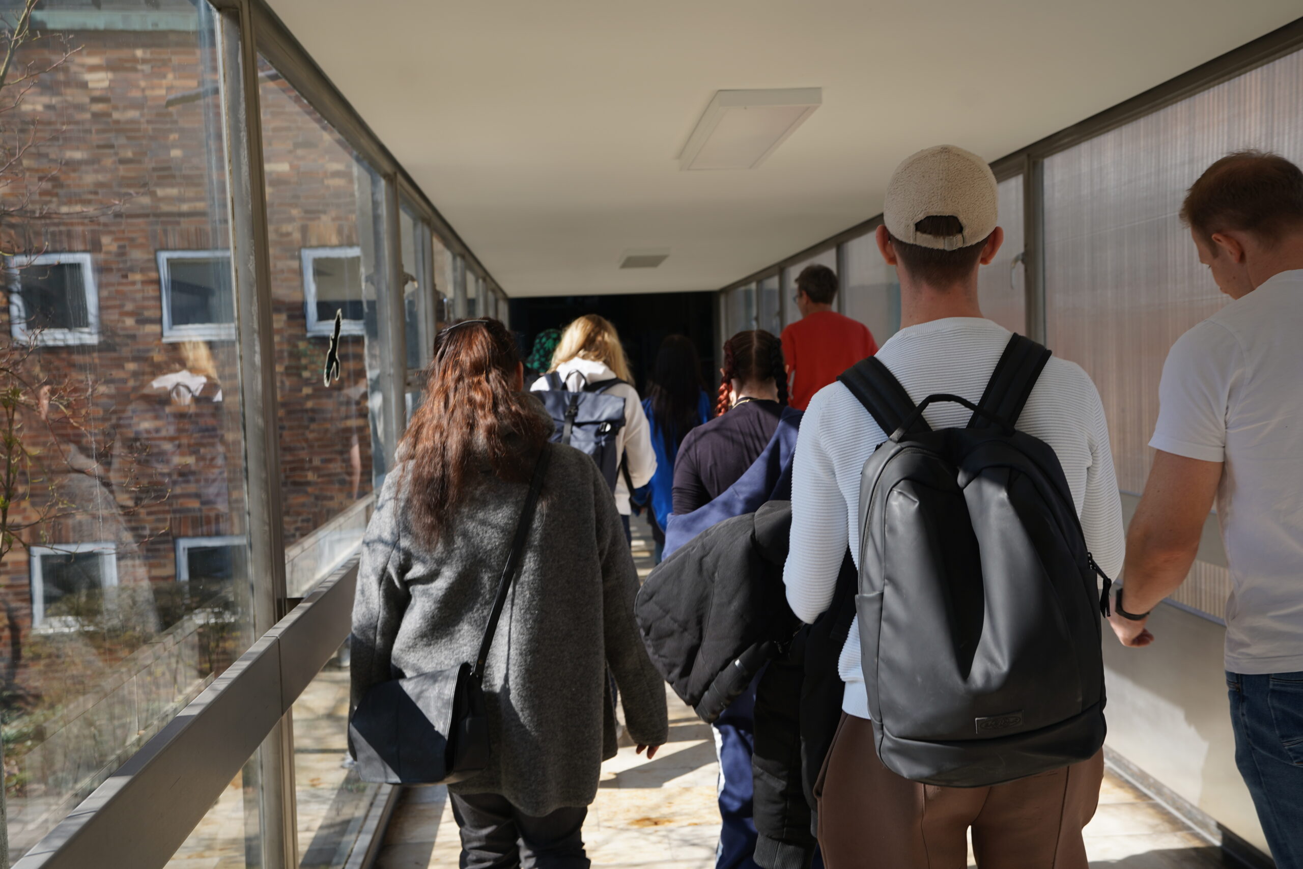 Eine Gruppe von Menschen mit Rucksäcken geht einen sonnenbeschienenen, geschlossenen Korridor mit Glaswänden auf dem Hochschul-Campus entlang und steuert auf einen Gebäudeeingang zu - vielleicht für einen Experimentiertag oder ein Studieninfo-Programm. Die Szene ist von hinten aufgenommen. at Hochschule Coburg
