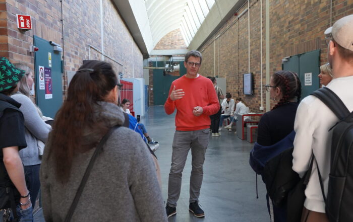 Ein Mann in einem roten Pullover spricht zu einer Gruppe von Menschen auf dem Hochschul-Campus, unter einer hohen, gewölbten Decke mit Backsteinwänden. Die Gruppe hört aufmerksam zu - einige tragen Rucksäcke - während des Studieninfo-Programms. at Hochschule Coburg