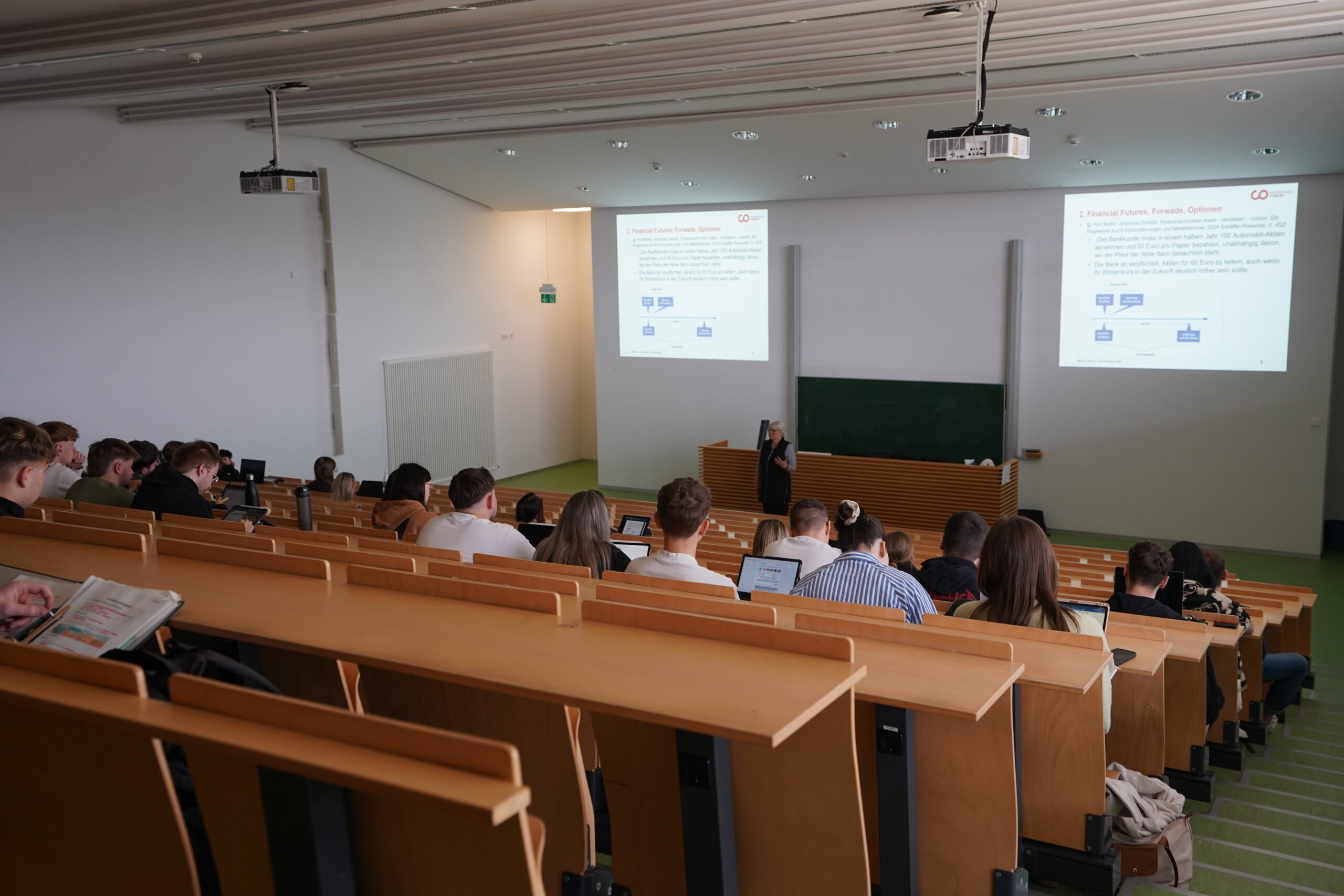 Studierende sitzen in einem holzbestuhlten Hörsaal auf dem Hochschul-Campus und schauen einem Dozenten bei einem Vortrag zu. Auf zwei Projektionsflächen werden Folien des Studieninfo-Programms im vorderen Teil des Hörsaals gezeigt. at Hochschule Coburg