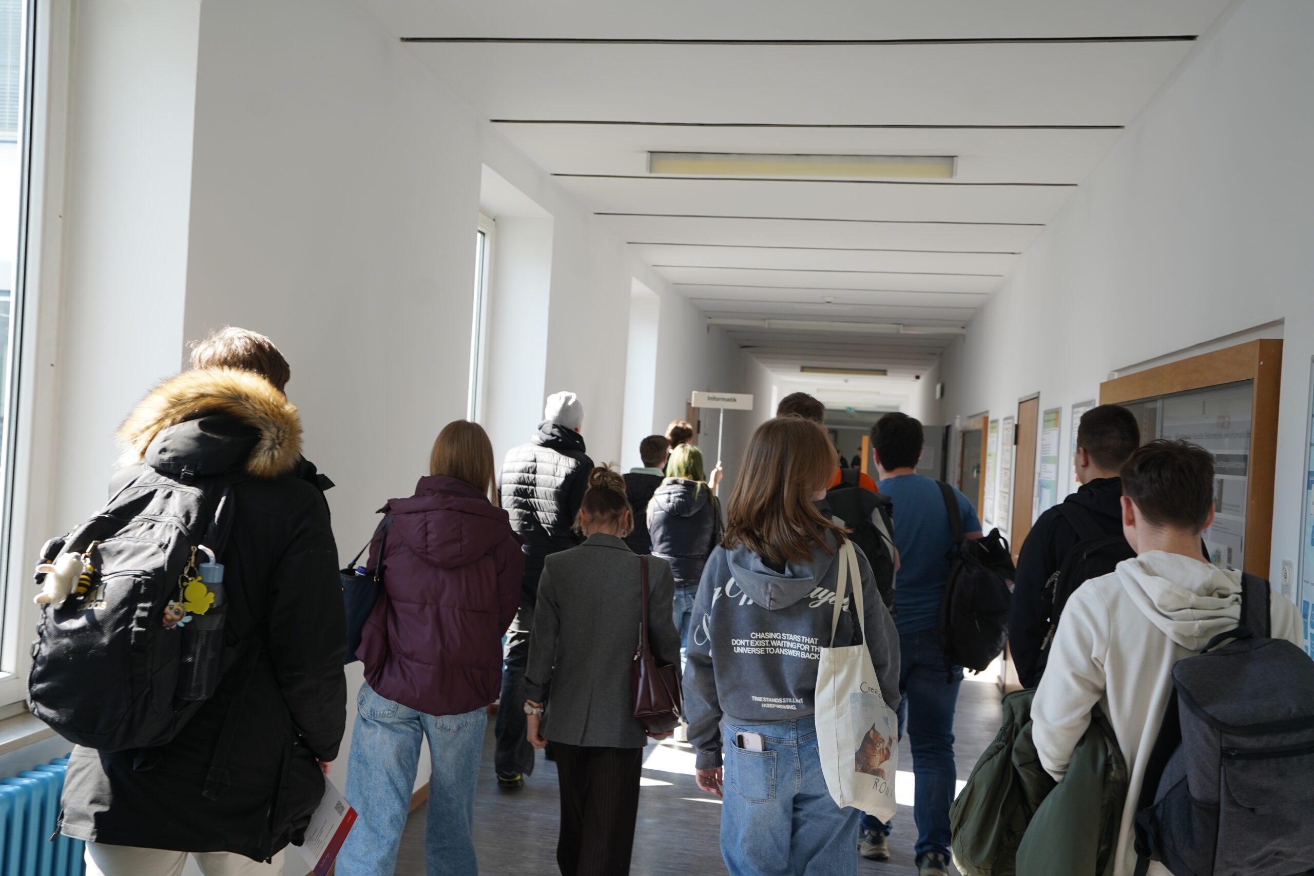 Eine Gruppe von Schülern mit Rucksäcken geht einen hellen Schulflur entlang, der durch das Sonnenlicht, das durch die Fenster auf der linken Seite fällt, erhellt wird. Sie sind auf dem Weg zu einem Experimentiertag im Rahmen des Studieninfo-Programms an Thüringer Schulen. at Hochschule Coburg