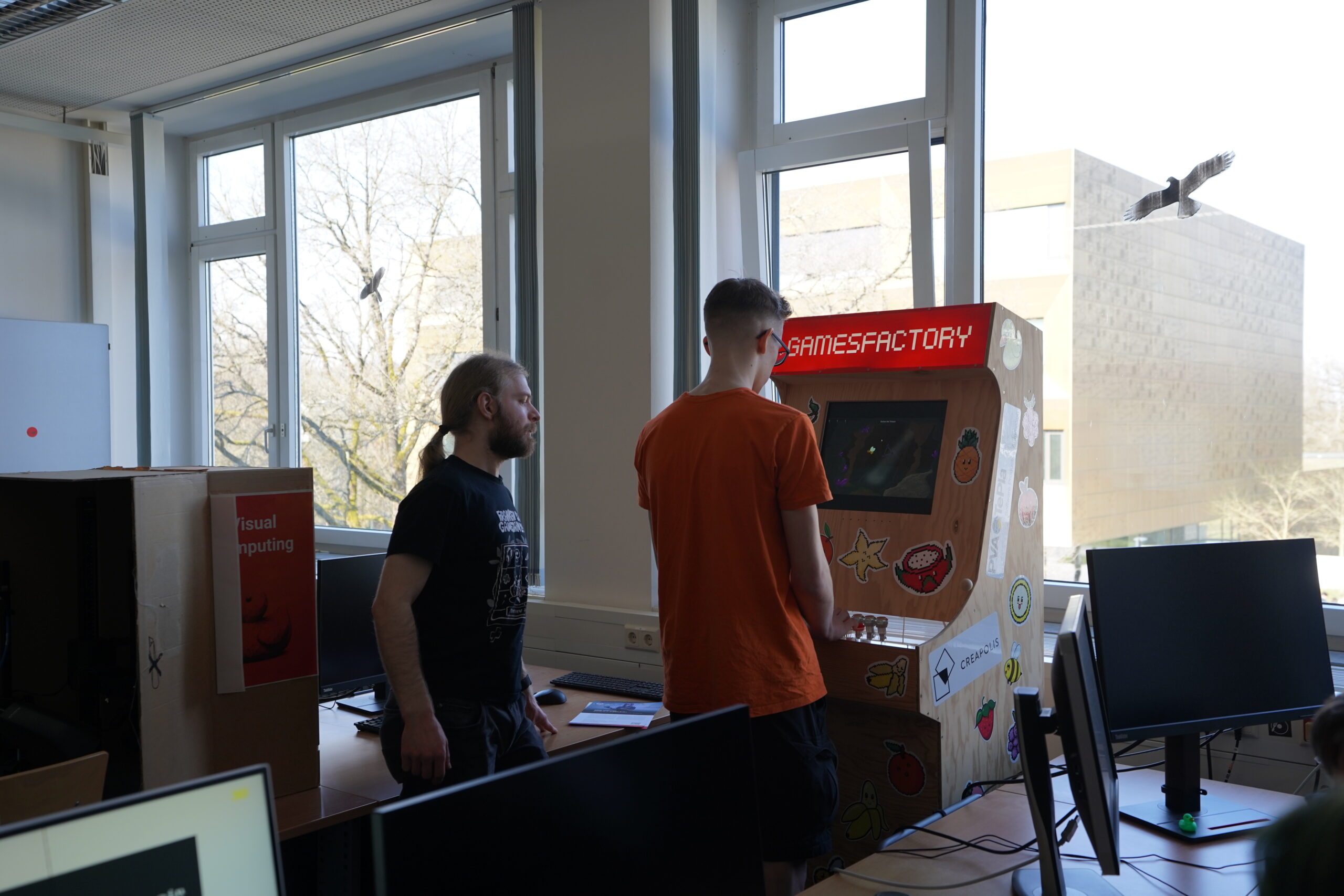 Zwei Personen stehen neben einem bunten "GAMESFACTORY"-Automaten in einem sonnendurchfluteten Raum mit großen Fenstern auf dem Hochschul-Campus. Eine Person schaut auf den Bildschirm, die andere auf den Automaten. In der Nähe befinden sich Computermonitore und Schreibtische. at Hochschule Coburg
