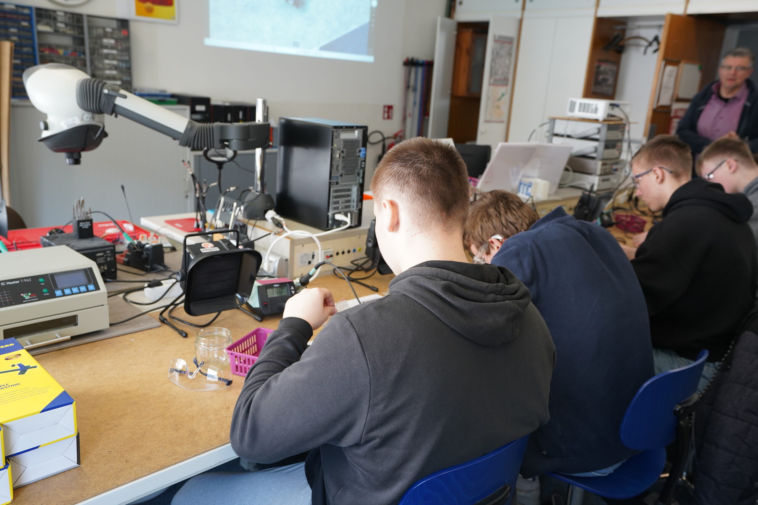 Mehrere Personen sitzen an einem Tisch auf dem Hochschul-Campus und arbeiten mit elektronischen Bauteilen und Geräten. Auf den Tischen sind Werkzeuge, Drähte und Geräte für einen Experimentiertag ausgebreitet. Im Hintergrund steht eine Person neben Regalen und einer Projektionsfläche. at Hochschule Coburg
