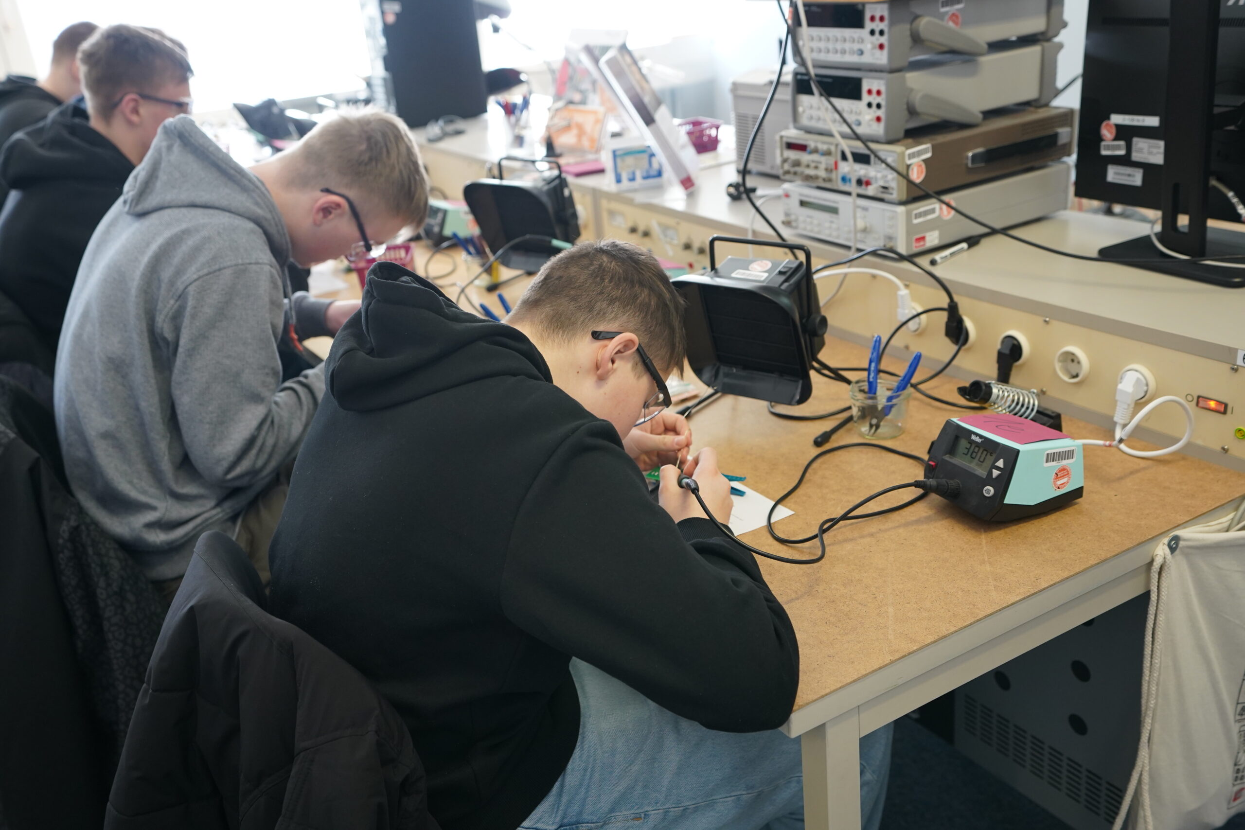 Zwei Personen in Kapuzenpullis sitzen während eines Experimentiertages auf dem Hochschul-Campus an einer unübersichtlichen Werkbank mit Lötkolben, umgeben von elektronischen Geräten und Kabeln. at Hochschule Coburg