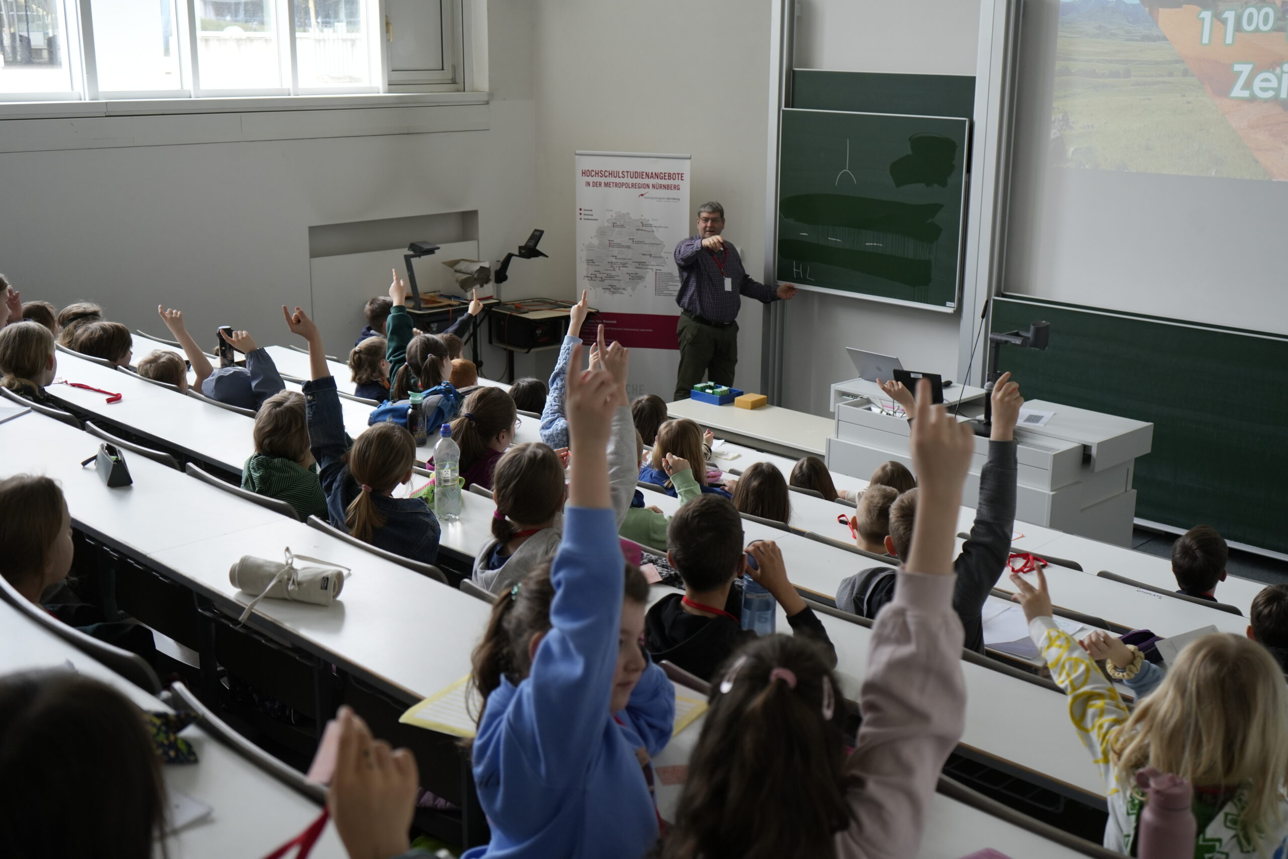 Ein Lehrer steht vorne im Hörsaal der KinderUni und spricht zu einer Gruppe von Studierenden. Viele sitzen mit erhobenen Händen an den Tischen in Reihen und wollen eine Frage zum Thema Falschgeld beantworten. Eine Projektionsfläche und Kreidetafeln sind zu sehen. at Hochschule Coburg