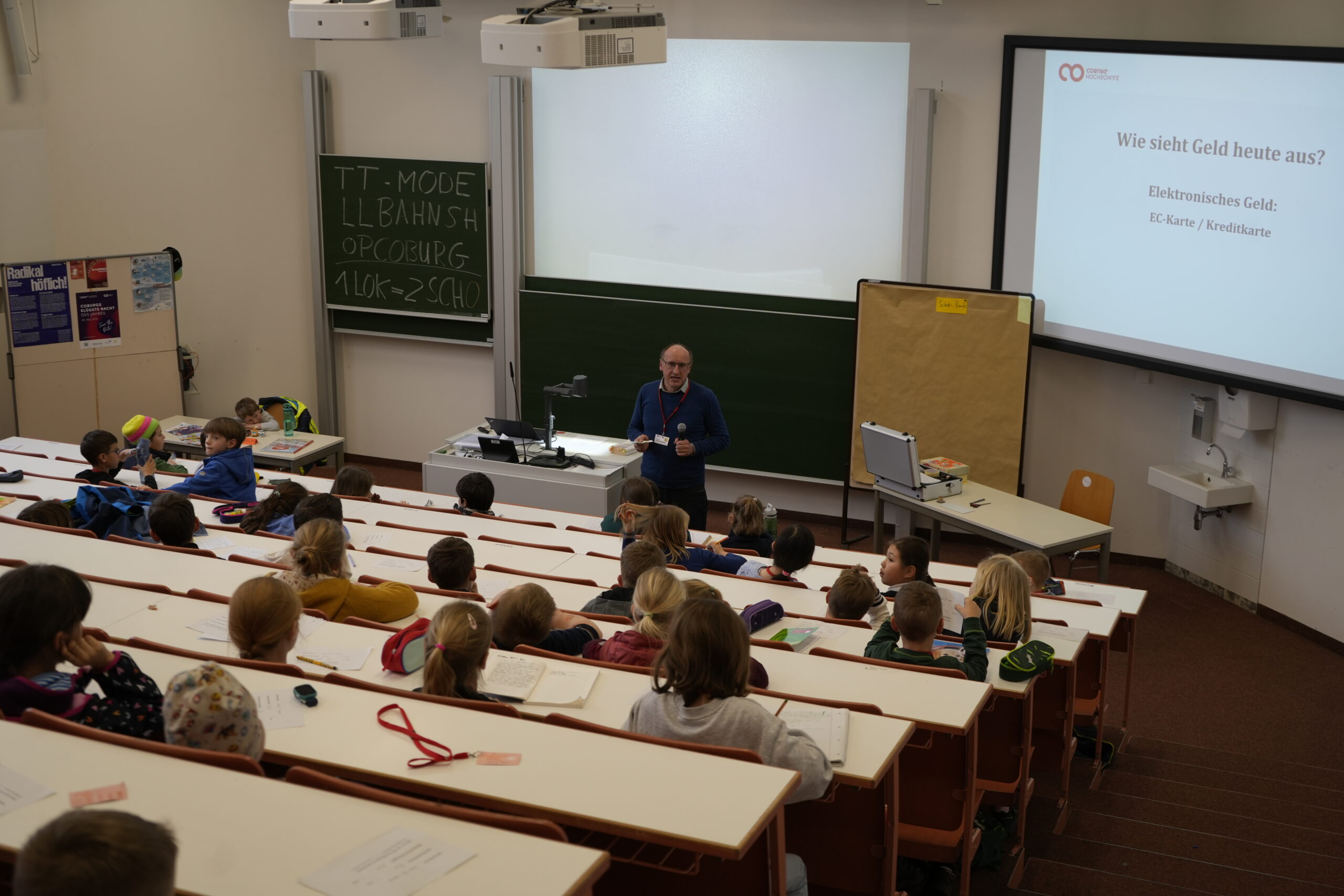 Ein Dozent steht vorne in einem Klassenzimmer der KinderUni und spricht zu kleinen Kindern, die in Reihen sitzen. Ein Projektor zeigt eine Folie über elektronisches Geld und KI, während eine Kreidetafel mit Schrift gefüllt ist. at Hochschule Coburg