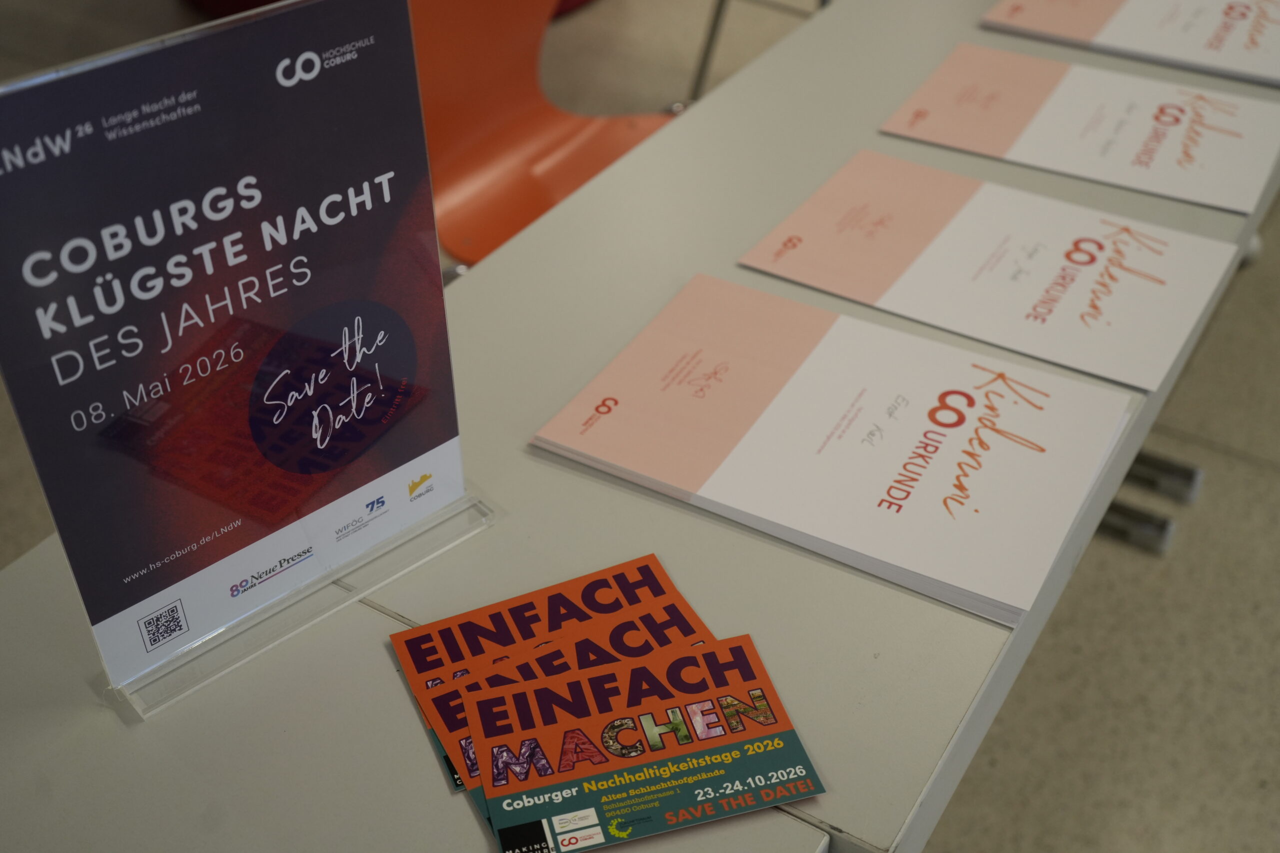 Auf einem Tisch liegen Veranstaltungsflyer, Urkunden und Postkarten für "Coburgs klügste Nacht des Jahres" am 8. Mai 2026 aus. Auf einem Schild steht "Save the Date", bunte Postkarten weisen auf Nachhaltigkeitstage und Themen wie KinderUni und KI hin. at Hochschule Coburg