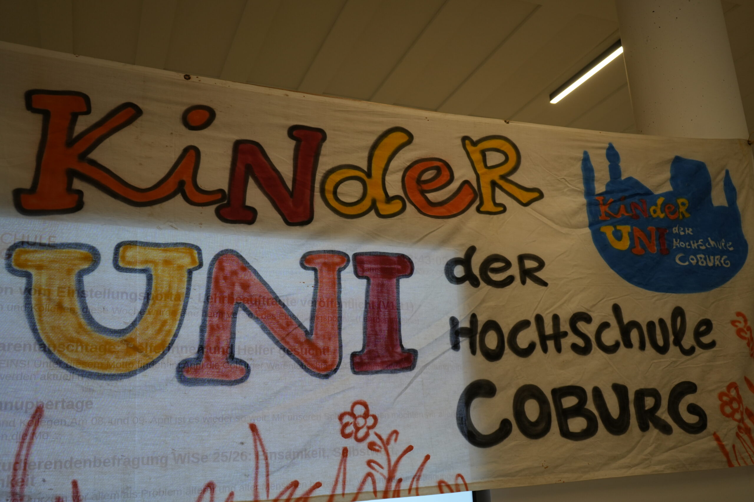 Auf einem farbenfrohen Banner steht in verspielter Schrift "Kinder UNI der Hochschule Coburg" und ein blaues KinderUni-Logo mit dem Schriftzug "Kinder UNI der Hochschule Coburg" und einer Stadtsilhouette. Orangefarbene Blumen zieren den Boden. at Hochschule Coburg