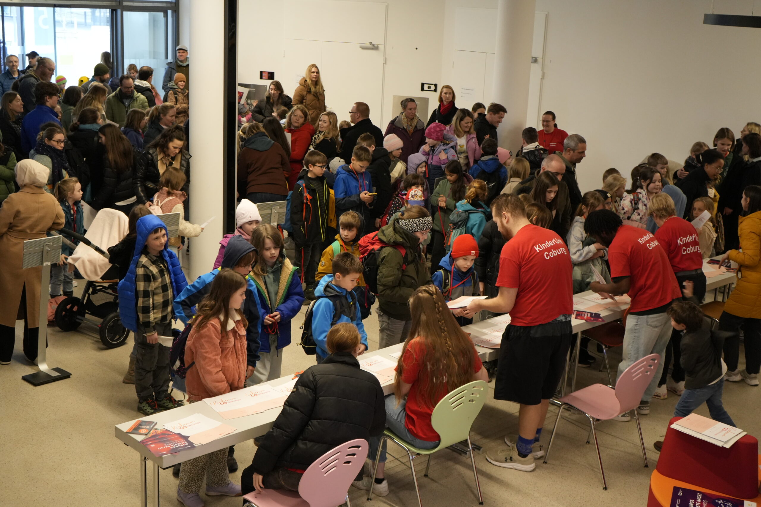 Eine große Gruppe von Kindern und Erwachsenen wartet in der Schlange am Anmeldeschalter der KinderUni, der von Mitarbeitern in roten Hemden in einem hellen Innenraum besetzt ist. Die Kinder tragen bunte Jacken und Hüte; die Atmosphäre ist geschäftig und lebhaft. at Hochschule Coburg