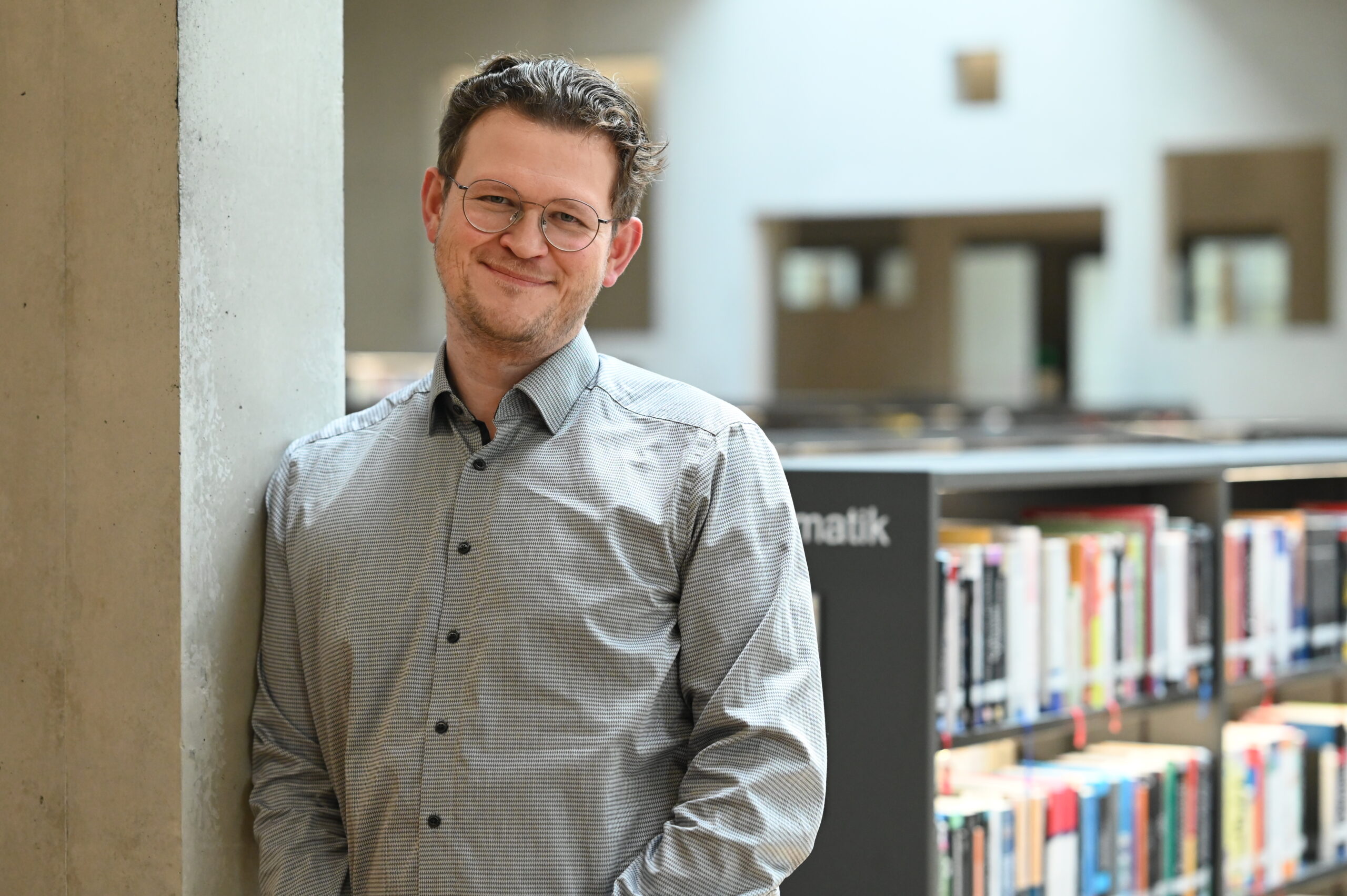 Ein Mann mit Brille und zugeknöpftem Hemd lehnt lächelnd an einer Säule in einer Bibliothek mit Bücherregalen und verschwommenen Büchern im Hintergrund. at Hochschule Coburg