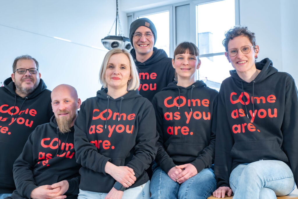 Sechs Personen sitzen und stehen in einem Raum zusammen, lächeln und tragen passende schwarze Kapuzenpullis mit der Aufschrift "Come as you are". Große Fenster und weiße Wände schaffen ein modernes Ambiente, perfekt für ein Treffen des Wirtschaftsinformatik-Teams. at Hochschule Coburg