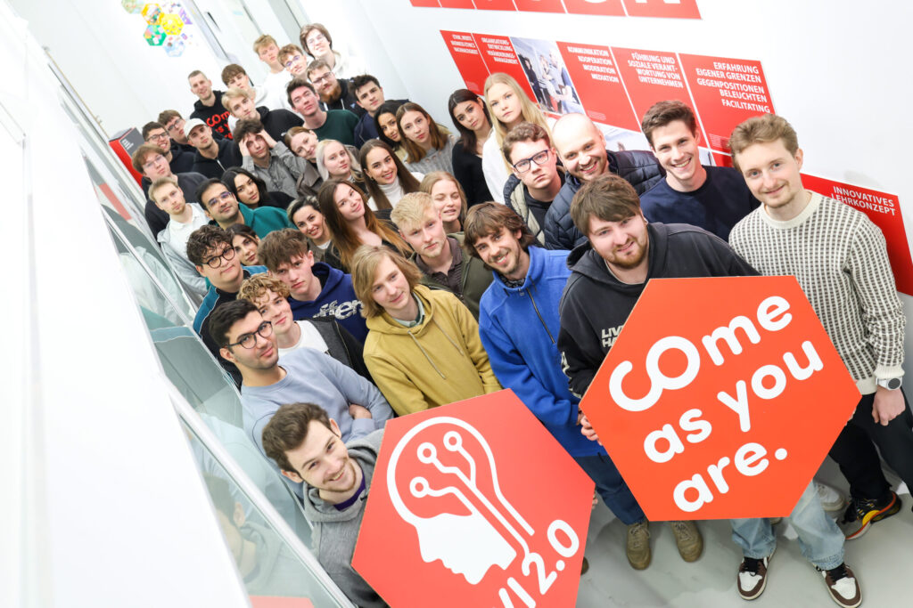 A large group of young adults stand together indoors, smiling and looking up at the camera. Two people in front hold red signs, one reading "Come as you are" and the other featuring a head with circuitry, reflecting interests in business administration and economy. at Hochschule Coburg