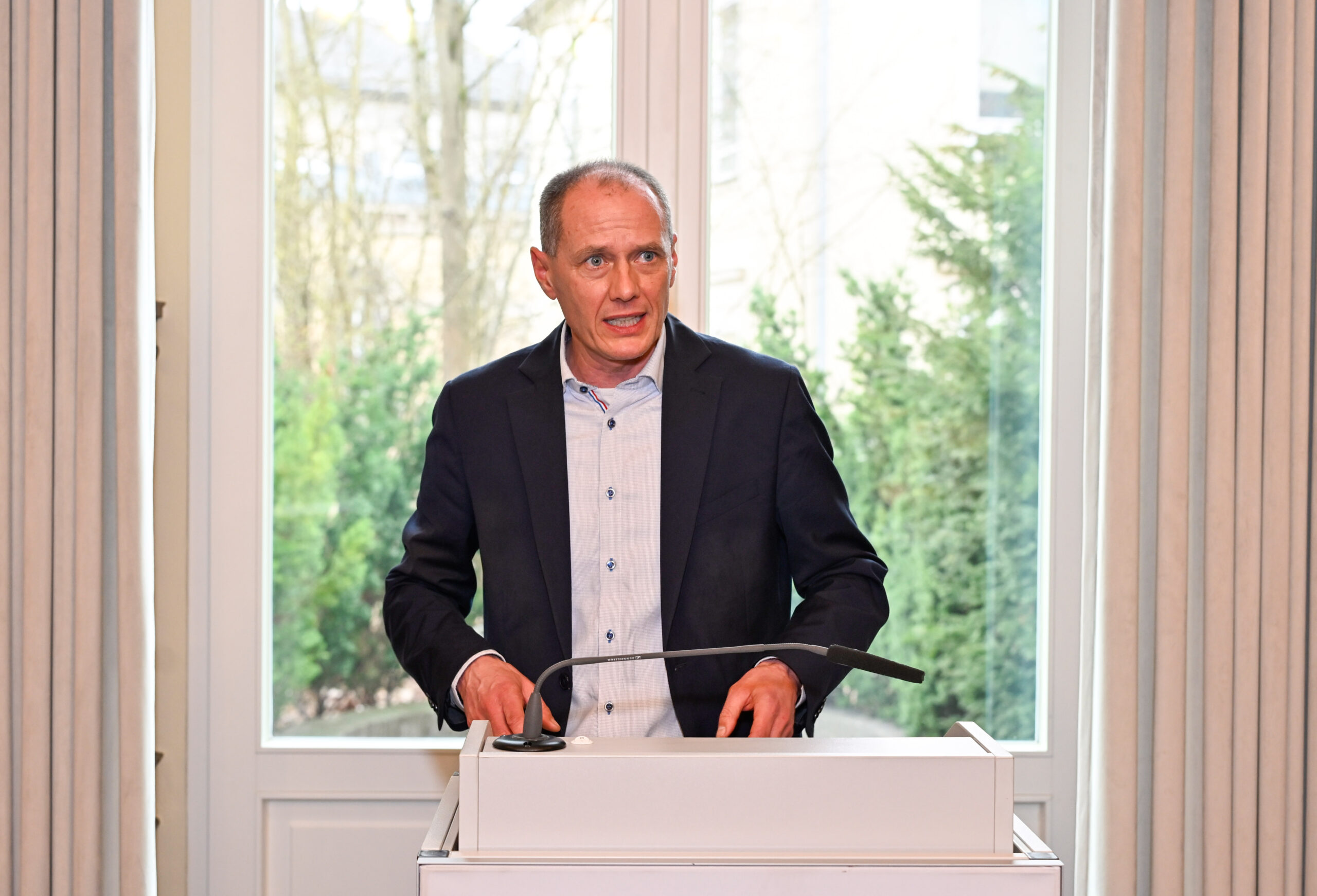 Ein Mann im Anzug steht hinter einem weißen Podium und spricht in einem hellen Raum mit großen Fenstern und beigen Vorhängen. Draußen sind grüne Bäume zu sehen, die auf eine Veranstaltung zum Wissenstransfer zwischen Hochschule und Wirtschaft hinweisen. at Hochschule Coburg