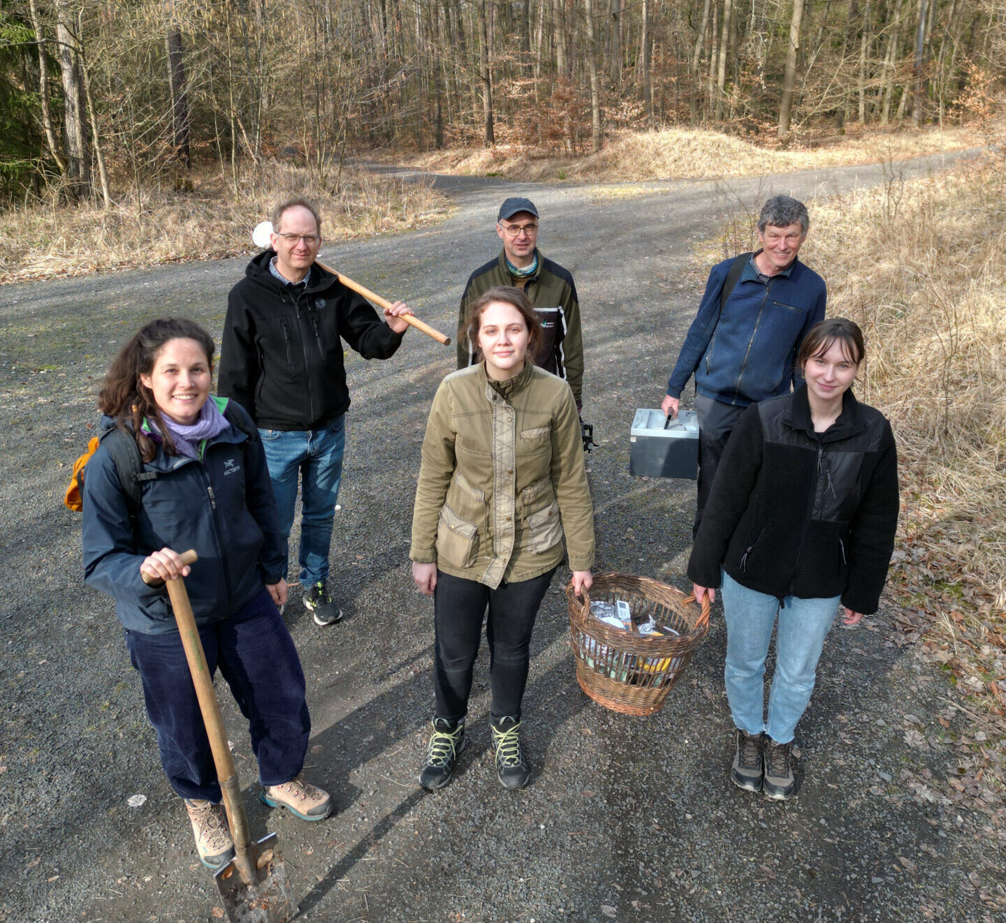 Fünf Personen stehen auf einem Kiesweg im Wald, halten Werkzeuge und einen Korb für ihr Forschungsprojekt in der Hand, tragen Outdoor-Kleidung und schauen an einem bewölkten Tag in die Kamera. at Hochschule Coburg