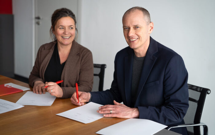 Zwei Personen sitzen an einem Tisch, lächelnd und mit roten Stiften in der Hand, mit Dokumenten vor sich, als befänden sie sich in einer wissenschaftlichen Kooperation oder beim Unterzeichnen sozialer Vereinbarungen in einem modernen Büro. at Hochschule Coburg