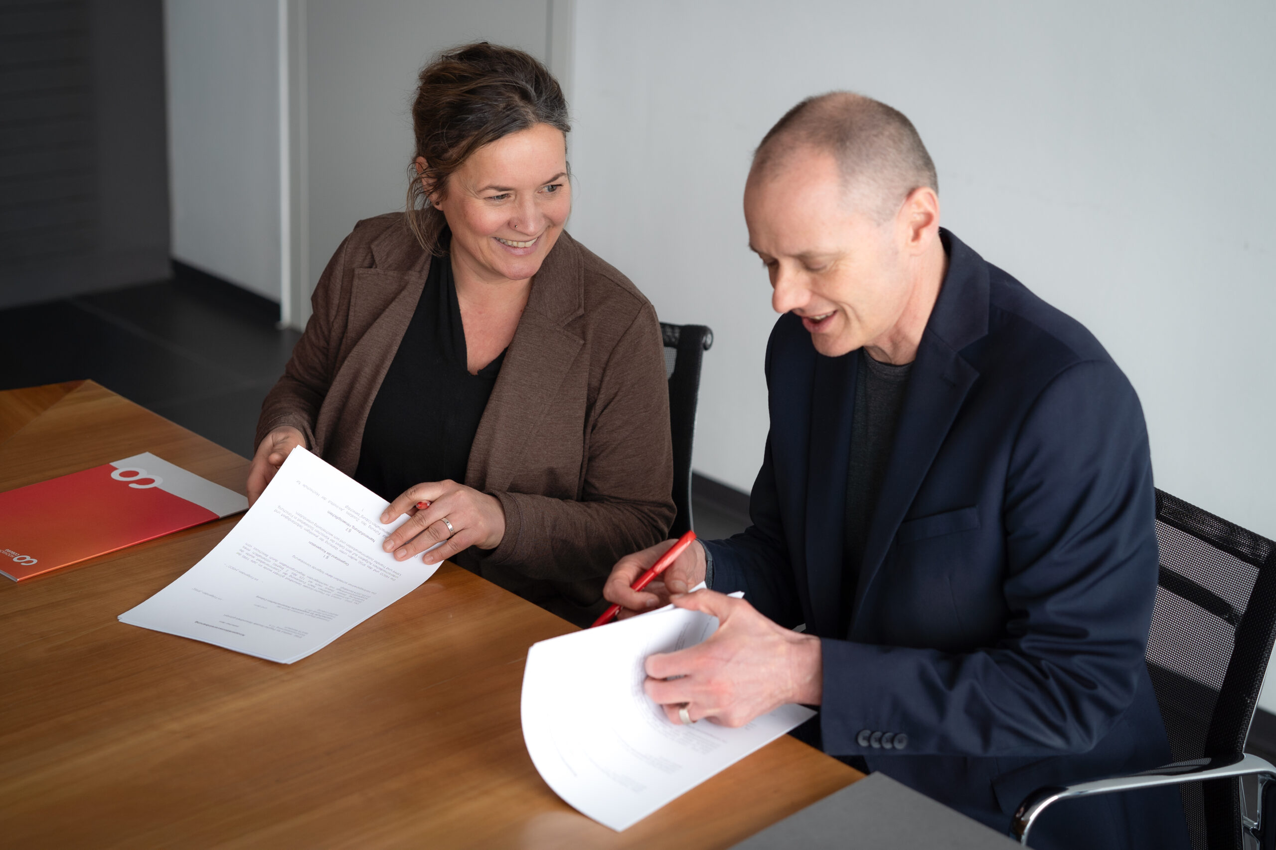 Zwei Personen sitzen an einem Tisch, lächeln und halten Papiere in der Hand. Der Mann unterschreibt ein Dokument mit einem roten Stift, während die Frau zuschaut. Beide scheinen in eine wissenschaftliche Kooperation verwickelt zu sein. at Hochschule Coburg