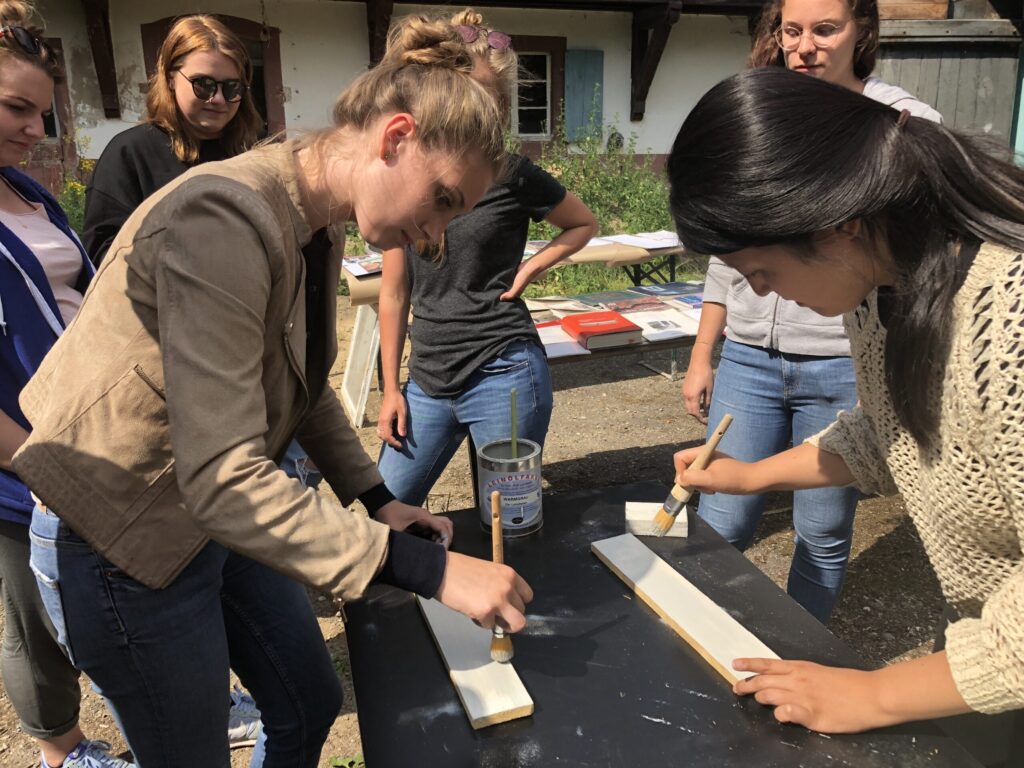 Eine Gruppe von Frauen im Freien sieht zu, wie zwei Frauen Holzbretter auf einem schwarzen Tisch bemalen und dabei einen praktischen Design-Workshop durchführen. Im Hintergrund sind Malutensilien und andere Materialien zu sehen, die auf eine kreative und gemeinschaftliche Atmosphäre hindeuten. at Hochschule Coburg