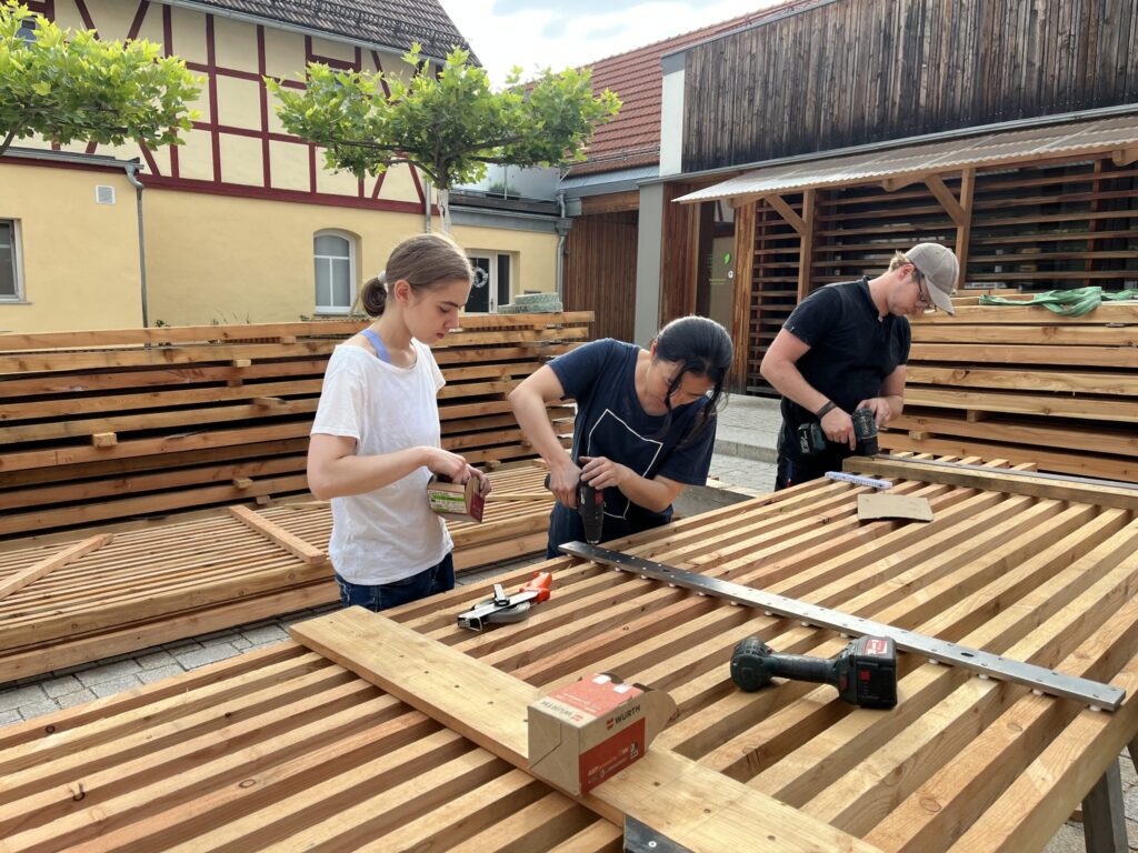 Drei Personen arbeiten gemeinsam im Freien und montieren große Holzplatten mit Werkzeugen und Schrauben. Sie tragen zu einem innovativen Designprojekt in Archiektur bei, umgeben von Holzstapeln, mit Gebäuden und Bäumen im Hintergrund. at Hochschule Coburg