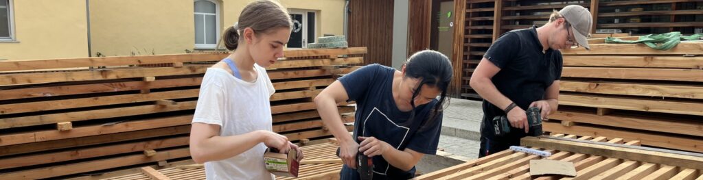Drei Personen arbeiten gemeinsam im Freien und montieren große Holzplatten mit Werkzeugen und Schrauben. Sie tragen zu einem innovativen Designprojekt in Archiektur bei, umgeben von Holzstapeln, mit Gebäuden und Bäumen im Hintergrund. at Hochschule Coburg