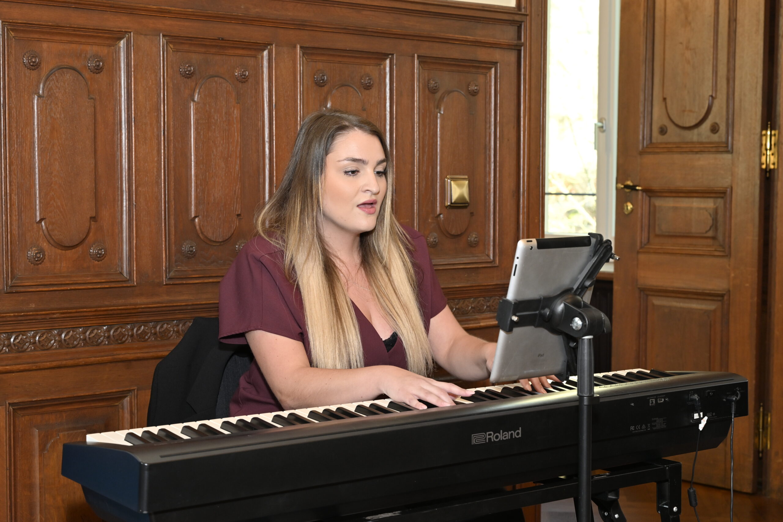Eine Frau mit langen blonden Haaren spielt ein elektrisches Keyboard und singt, während sie von einem Tablet auf einem Ständer abliest, in einem Raum mit verzierter Holzvertäfelung - eine inspirierende Szene für Wissenstransfer zwischen Hochschule und Wirtschaft. at Hochschule Coburg