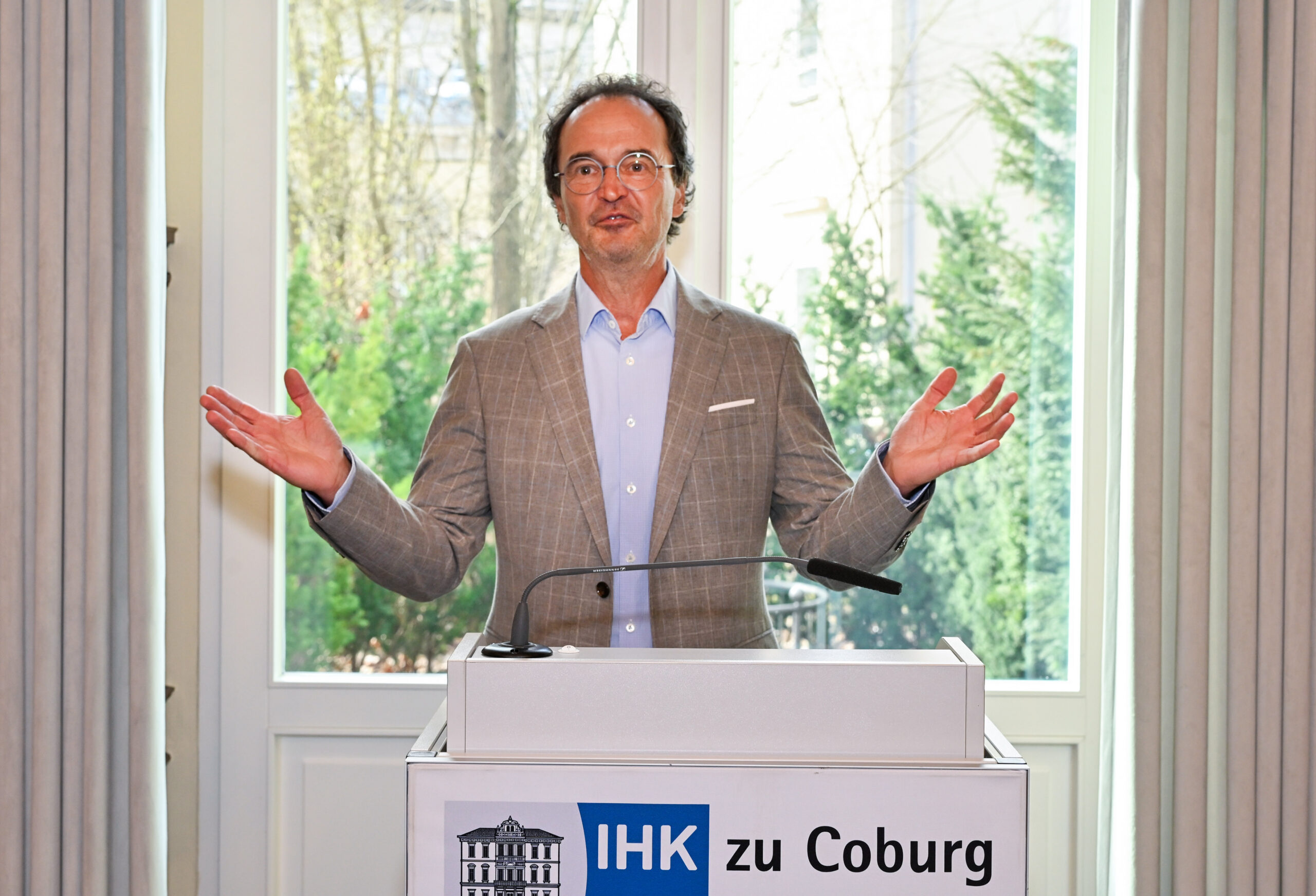 Ein Mann in einer Anzugsjacke steht an einem Podium mit einem Mikrofon und gestikuliert mit offenen Armen. Auf dem Podium prangt ein Schild mit der Aufschrift "IHK zu Coburg". Große Fenster und Grünflächen sind zu sehen - ein Hinweis auf den Wissenstransfer zwischen Hochschule und Wirtschaft. at Hochschule Coburg