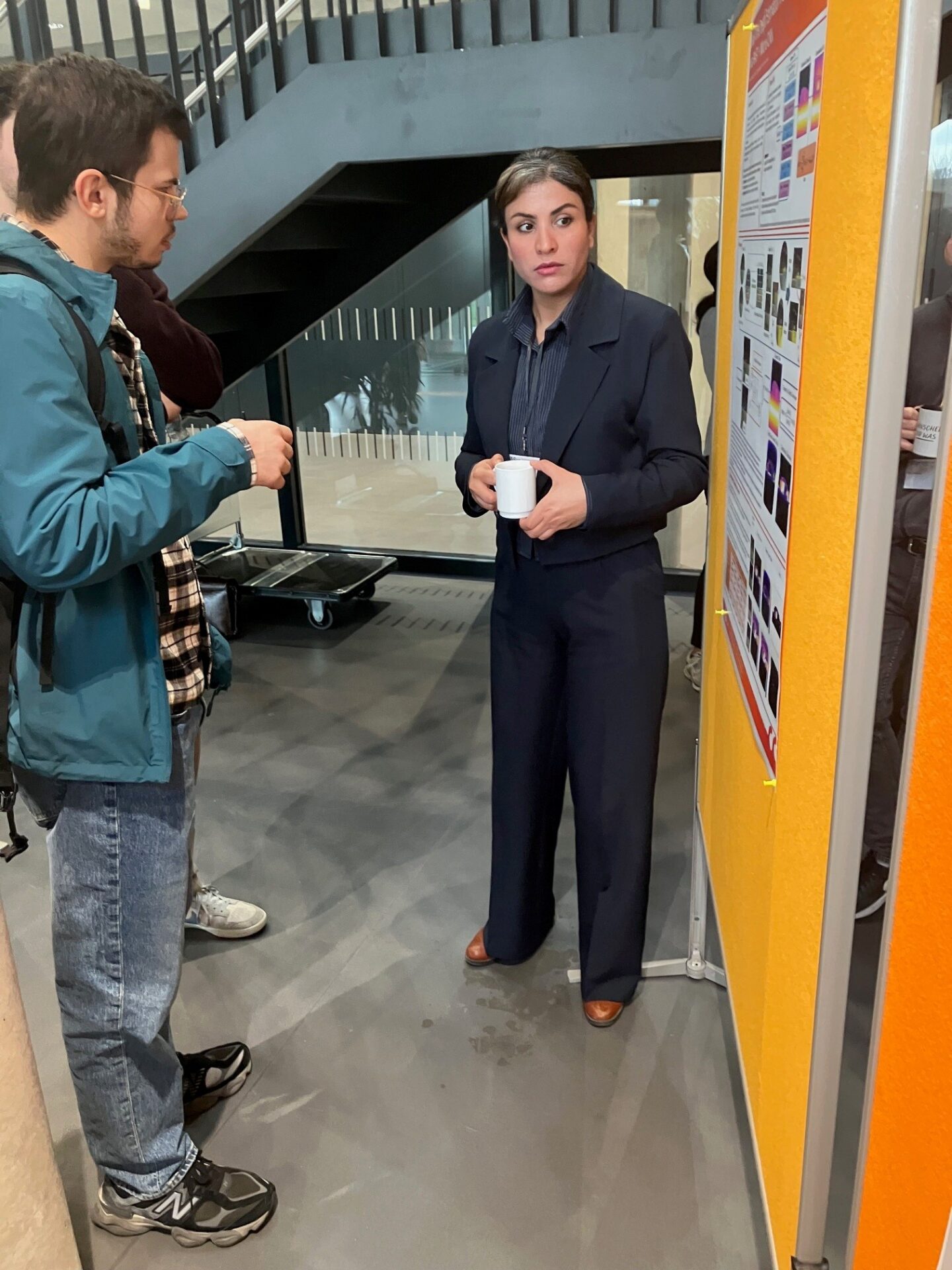 Eine Frau im dunklen Anzug hält einen weißen Becher in der Hand, während sie neben einem bunten akademischen Poster steht und sich mit einem Mann in Freizeitkleidung unterhält - ein inspirierender Austausch zwischen Doktoranden in einem Innenraum mit einer Treppe im Hintergrund. at Hochschule Coburg