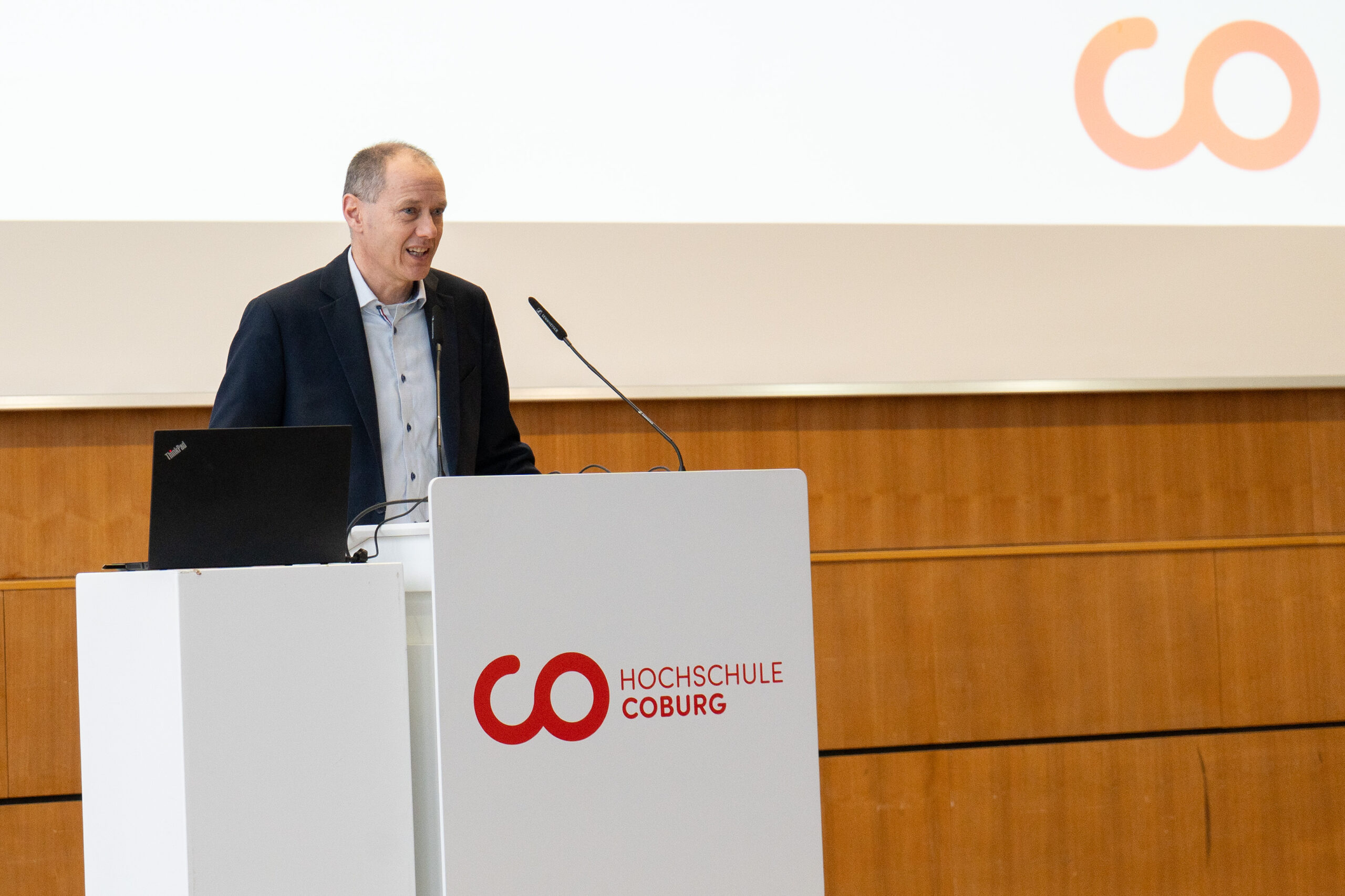 Ein Mann im Anzug spricht an einem Podium mit einem Mikrofon und einem Laptop. Das Logo der Hochschule Coburg ist in Rot auf Weiß zu sehen, was darauf hindeutet, dass es sich um eine formelle Veranstaltung handelt, bei der die Erstsemester zum Beginn ihres Studiums begrüßt werden. at Hochschule Coburg