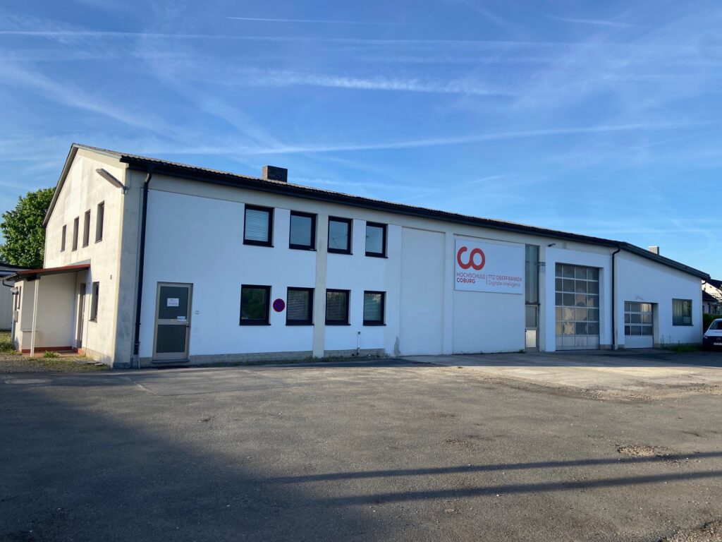 A white industrial building with multiple windows, a large garage door, and a "GO" sign on the front wall stands on a paved lot under a clear blue sky, near Coburg University. at Hochschule Coburg