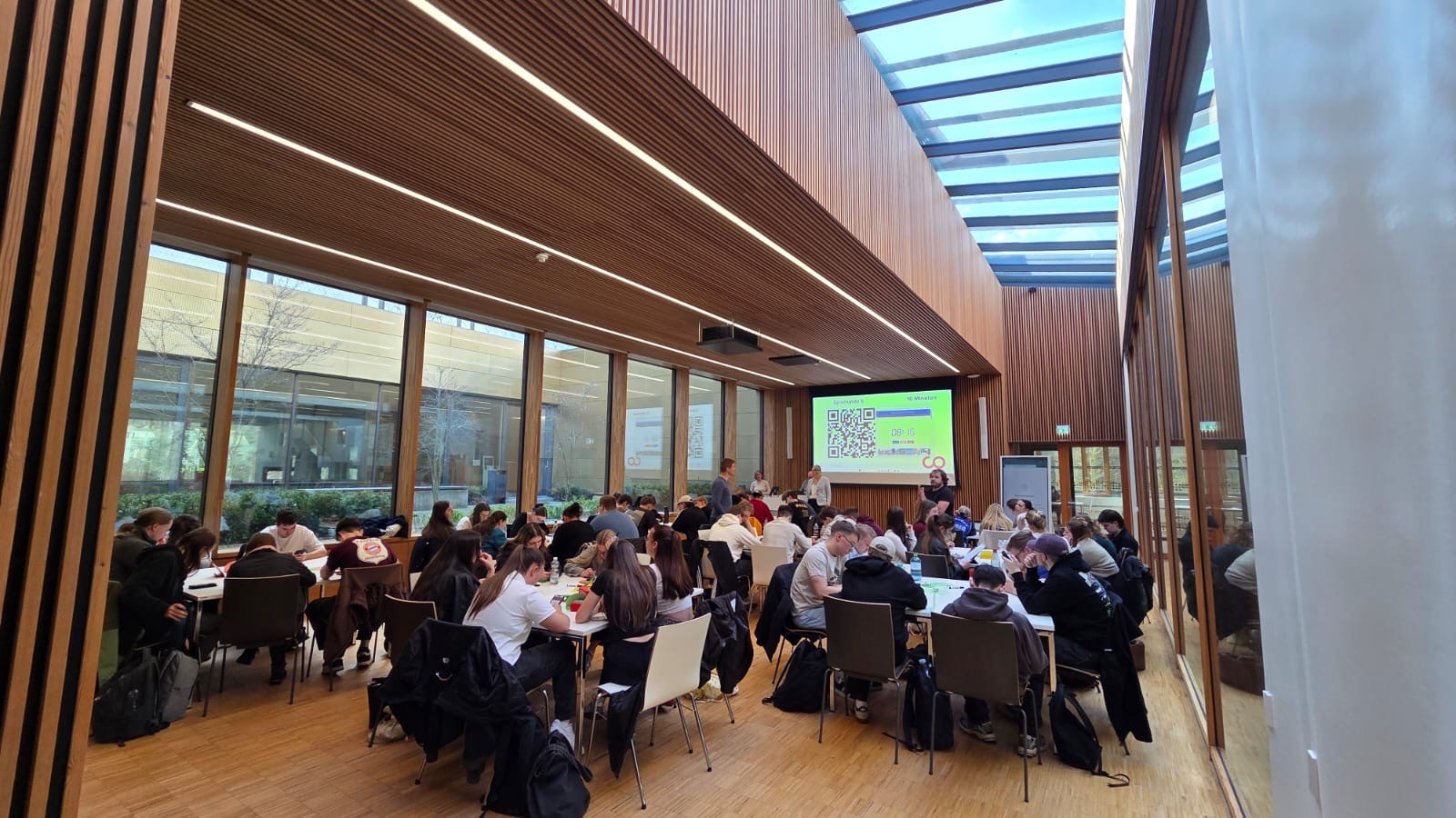 Eine große Gruppe versammelt sich an runden Tischen in einem modernen, holzgetäfelten Raum auf dem Hochschul-Campus. Ein Moderator steht vor einem Bildschirm mit einem QR-Code und Folien und führt durch das Studieninfo-Programm. at Hochschule Coburg