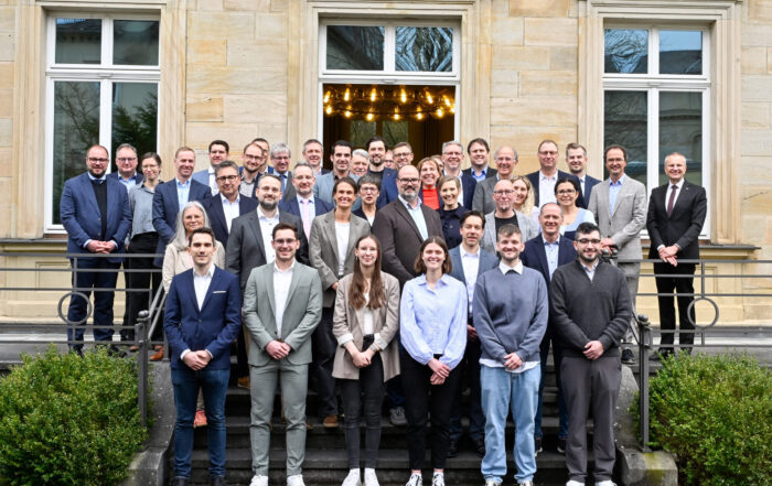 Eine große Gruppe professionell gekleideter Menschen posiert gemeinsam auf den Stufen vor einem stattlichen Hochschulgebäude mit großen Fenstern und Steinmauern und lächelt für ein Gruppenfoto. at Hochschule Coburg