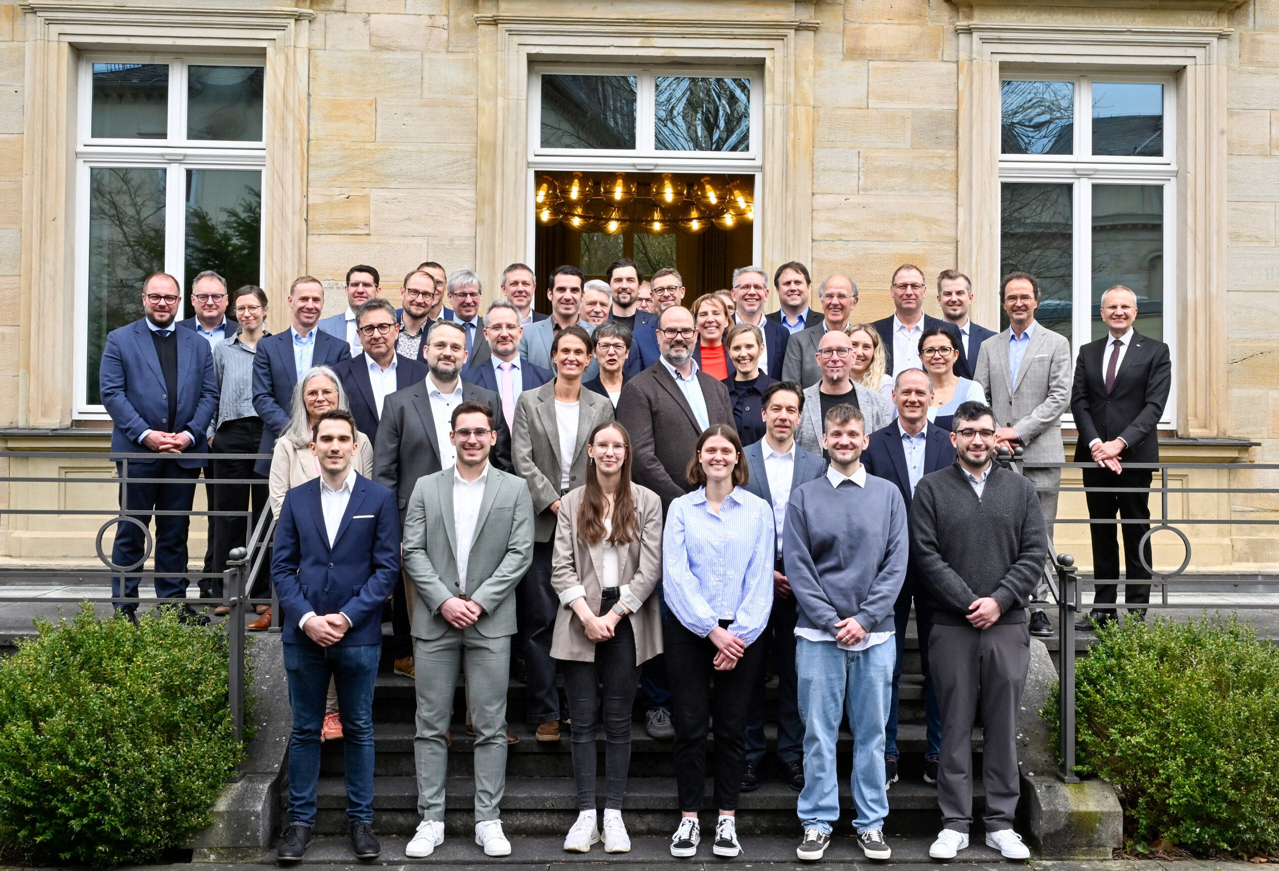 Eine große Gruppe professionell gekleideter Menschen posiert gemeinsam auf den Stufen vor einem stattlichen Hochschulgebäude mit großen Fenstern und Steinmauern und lächelt für ein Gruppenfoto. at Hochschule Coburg