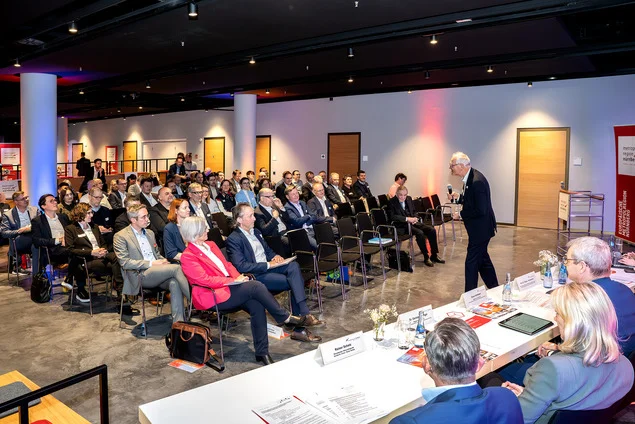Eine Gruppe von Menschen sitzt in Stuhlreihen bei einer Indoor-Konferenz in der Metropolregion und hört einem Redner an der Stirnseite zu. Ein Gremium des TTZ Oberfranken sitzt an einem Tisch, und auf den Tischen liegen Forschungsmaterialien. at Hochschule Coburg