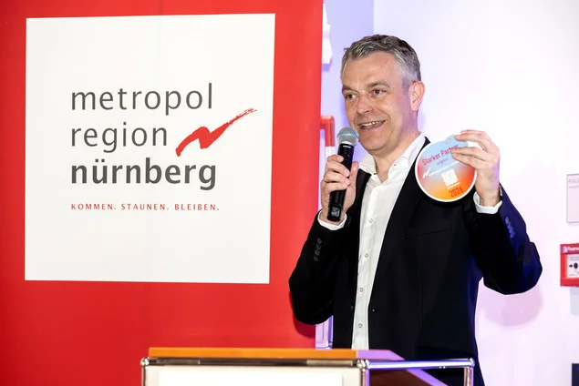 Ein Mann im Anzug spricht in ein Mikrofon und hält einen bunten, runden Gegenstand in der Hand. Er steht neben einem Schild mit der Aufschrift "metropol region nürnberg", das das Engagement der Metropolregion für Forschung und Innovation hervorhebt. at Hochschule Coburg