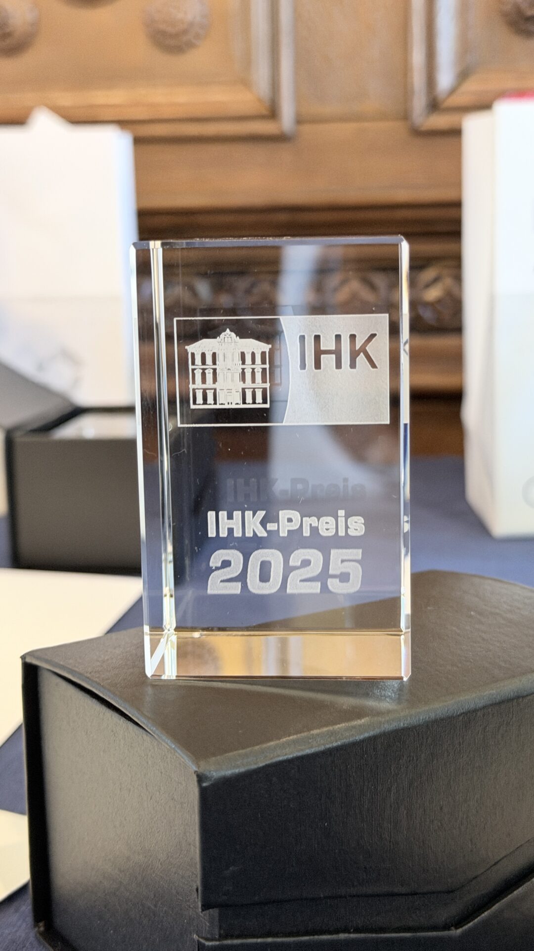 Ein klarer Glaspreis mit der Gravur "IHK" und einem Gebäudebild mit der Aufschrift "IHK-Preis 2025" steht auf einem schwarzen Sockel, der den Wissenstransfer zwischen Hochschule und Wirtschaft würdigt, mit unscharfen Kästen und einer dekorativen Holzverkleidung im Hintergrund. at Hochschule Coburg