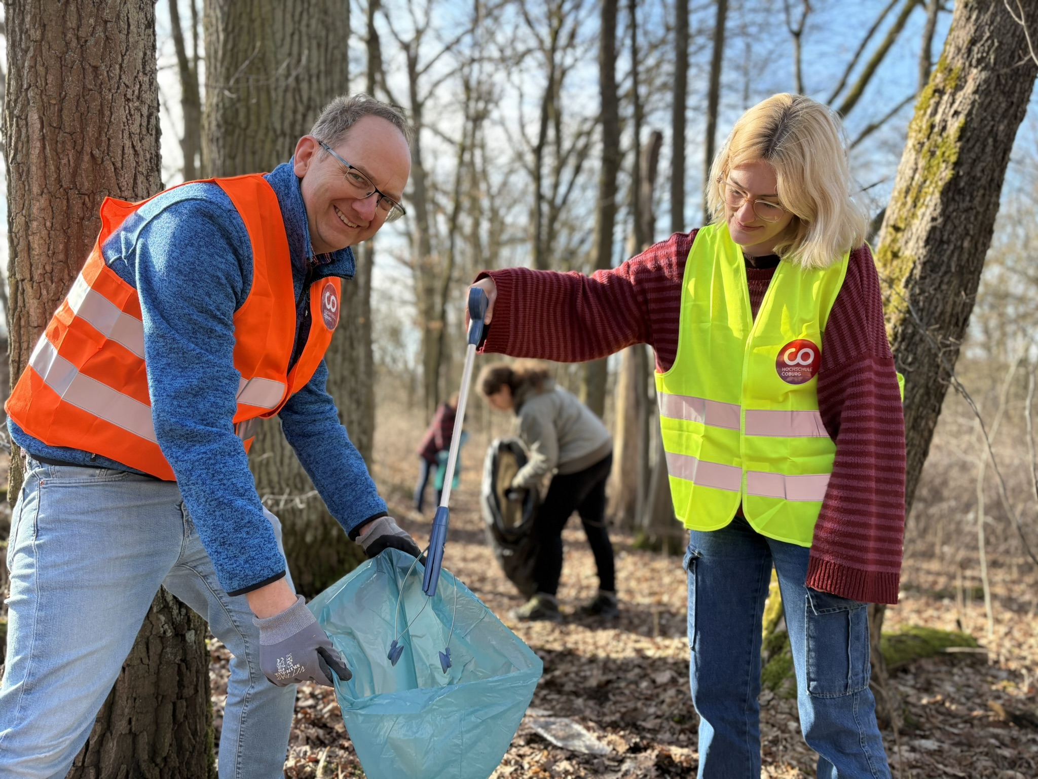 Zwei Personen in Sicherheitswesten sammeln in einem Wald Müll auf und füllen ihn in einen blauen Sack - eine Aktion, die den Anliegen unseres Kunststoff-Zeitalters entspricht. Andere Freiwillige räumen an diesem sonnigen Tag im Hintergrund auf. at Hochschule Coburg