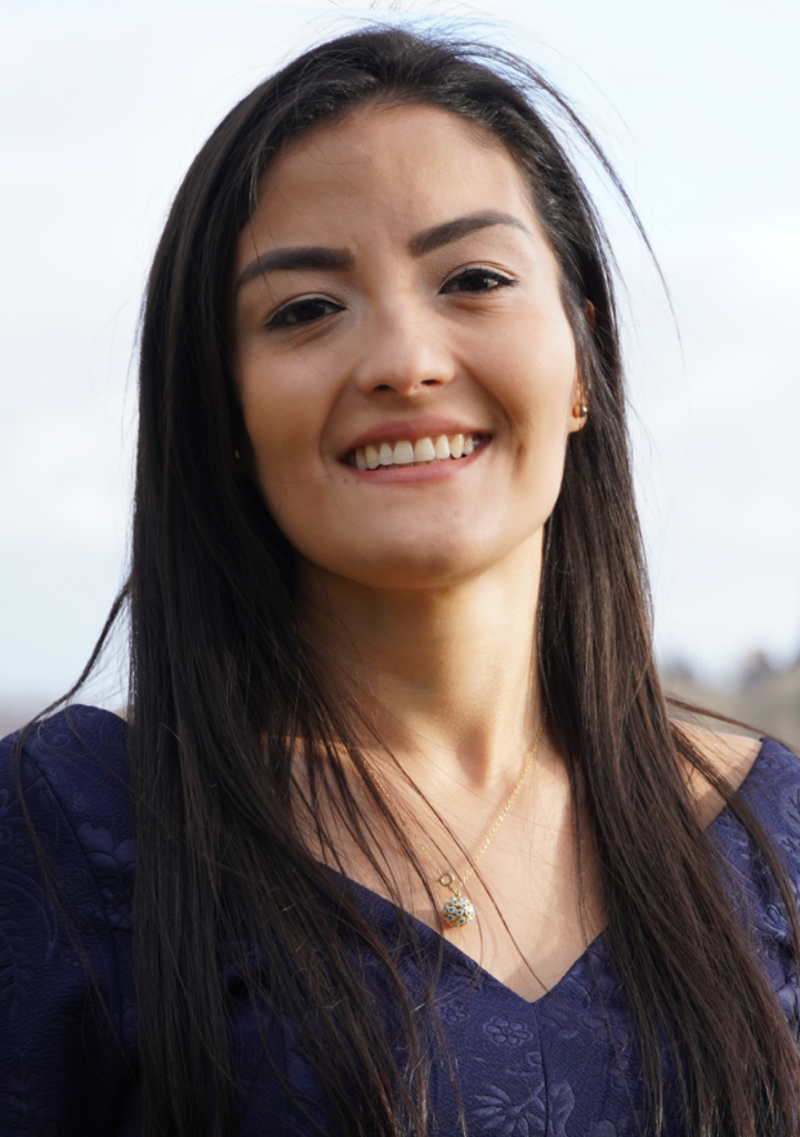 Eine Frau mit langen dunklen Haaren, die ein marineblaues Oberteil und eine zarte Halskette trägt, lächelt im Freien vor einem verschwommenen hellen Hintergrund. at Hochschule Coburg