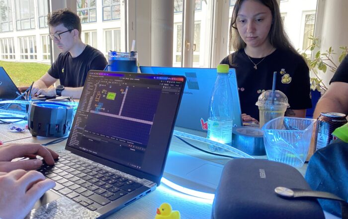 Drei Personen arbeiten an Laptops an einem Tisch mit Getränken, persönlichen Gegenständen und einer Gummiente. Ein Game Jam 2026-Projekt ist auf einem Bildschirm geöffnet, während das Sonnenlicht durch große Fenster strömt und Momente des Scheiterns in kreative Teamarbeit verwandelt. at Hochschule Coburg
