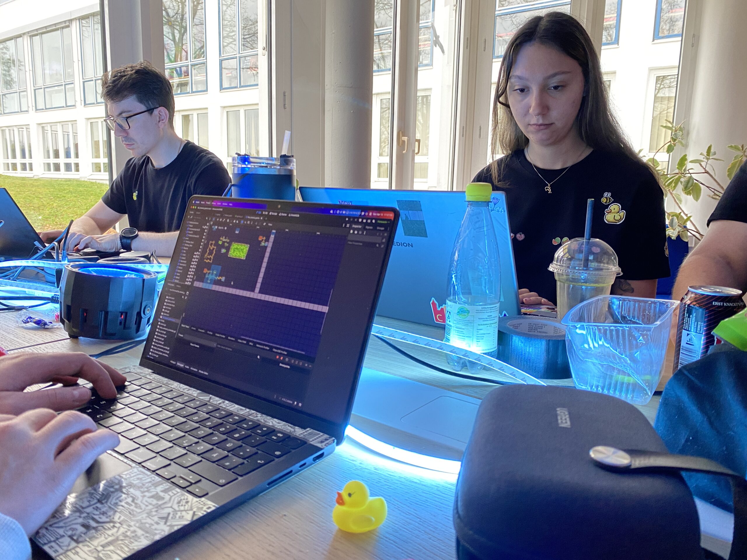 Drei Personen arbeiten an Laptops an einem Tisch mit Getränken, persönlichen Gegenständen und einer Gummiente. Ein Game Jam 2026-Projekt ist auf einem Bildschirm geöffnet, während das Sonnenlicht durch große Fenster strömt und Momente des Scheiterns in kreative Teamarbeit verwandelt. at Hochschule Coburg