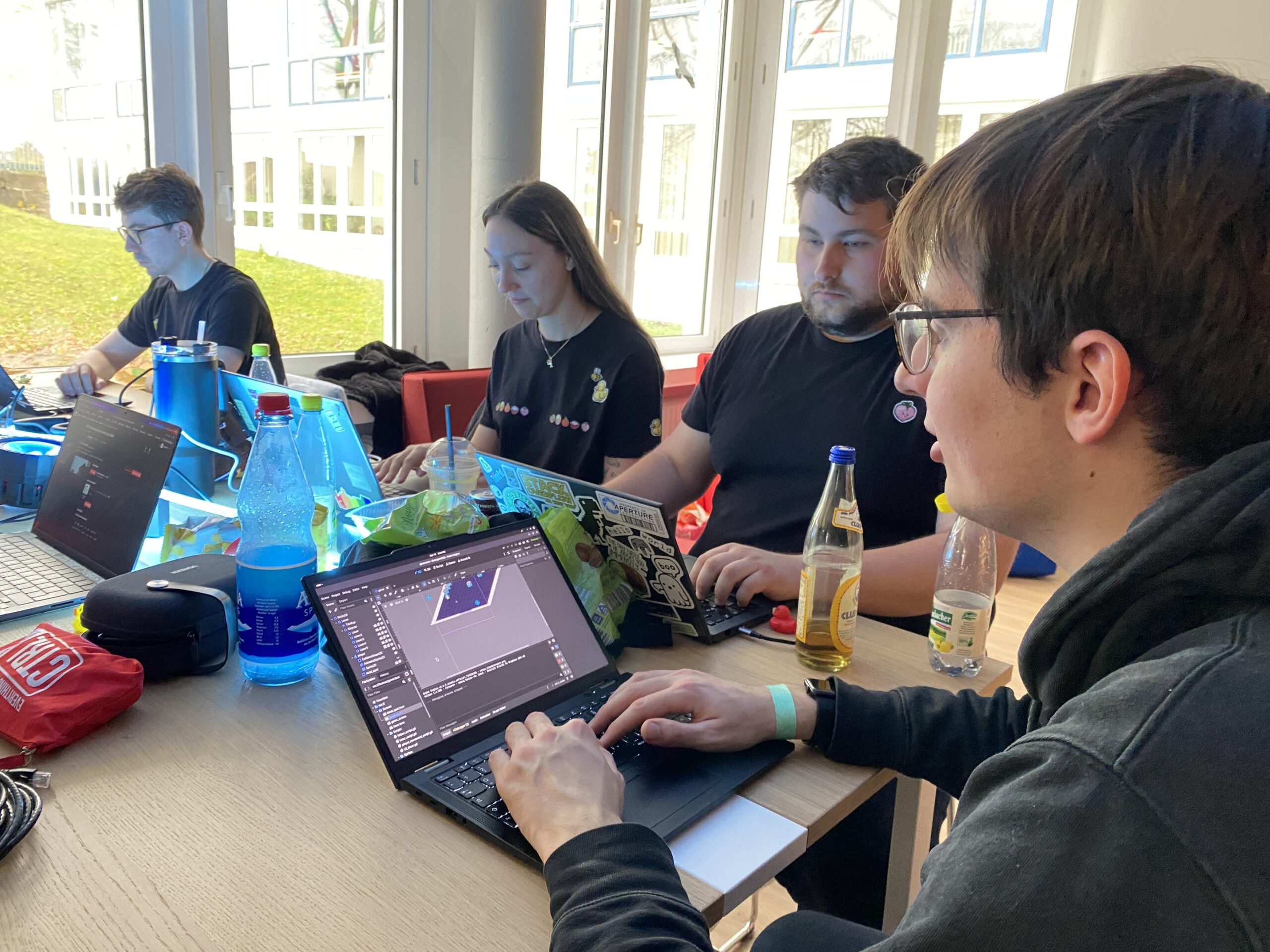 Vier Leute sitzen beim Game Jam 2026 an einem Tisch, arbeiten an Laptops und konzentrieren sich auf ihre Bildschirme. Wasserflaschen, Snacks und Getränke sind über den Tisch verstreut, während das Sonnenlicht durch die großen Fenster strömt - so wird aus dem Scheitern ein gemeinsamer Erfolg. at Hochschule Coburg