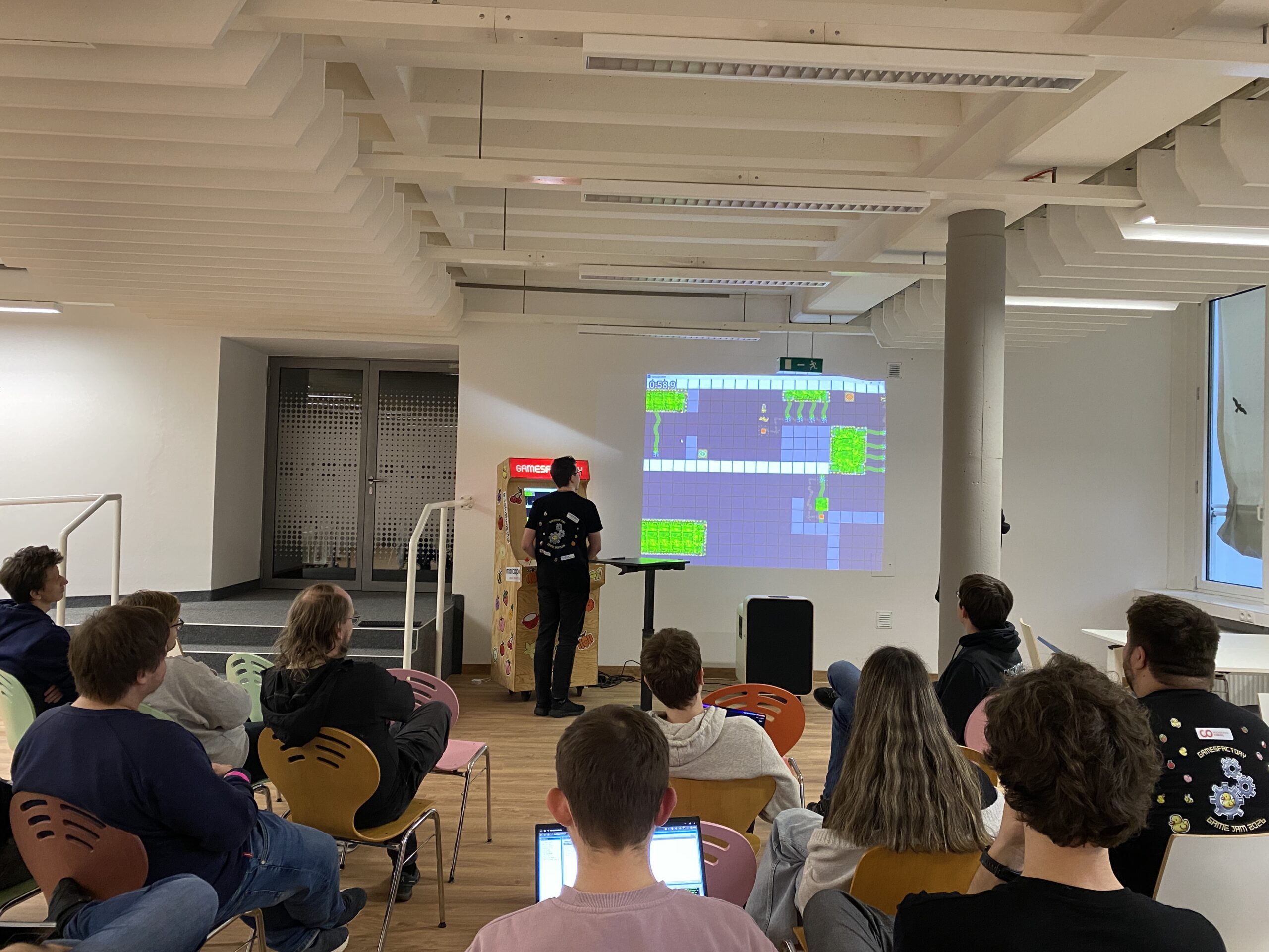 In einem hell erleuchteten Raum mit modernem Dekor und bunten Stühlen sitzt eine Gruppe von Menschen und sieht zu, wie eine Person ein Videospiel vorstellt, das während des Game Jam 2026 entwickelt wurde und an die Wand projiziert wird. at Hochschule Coburg