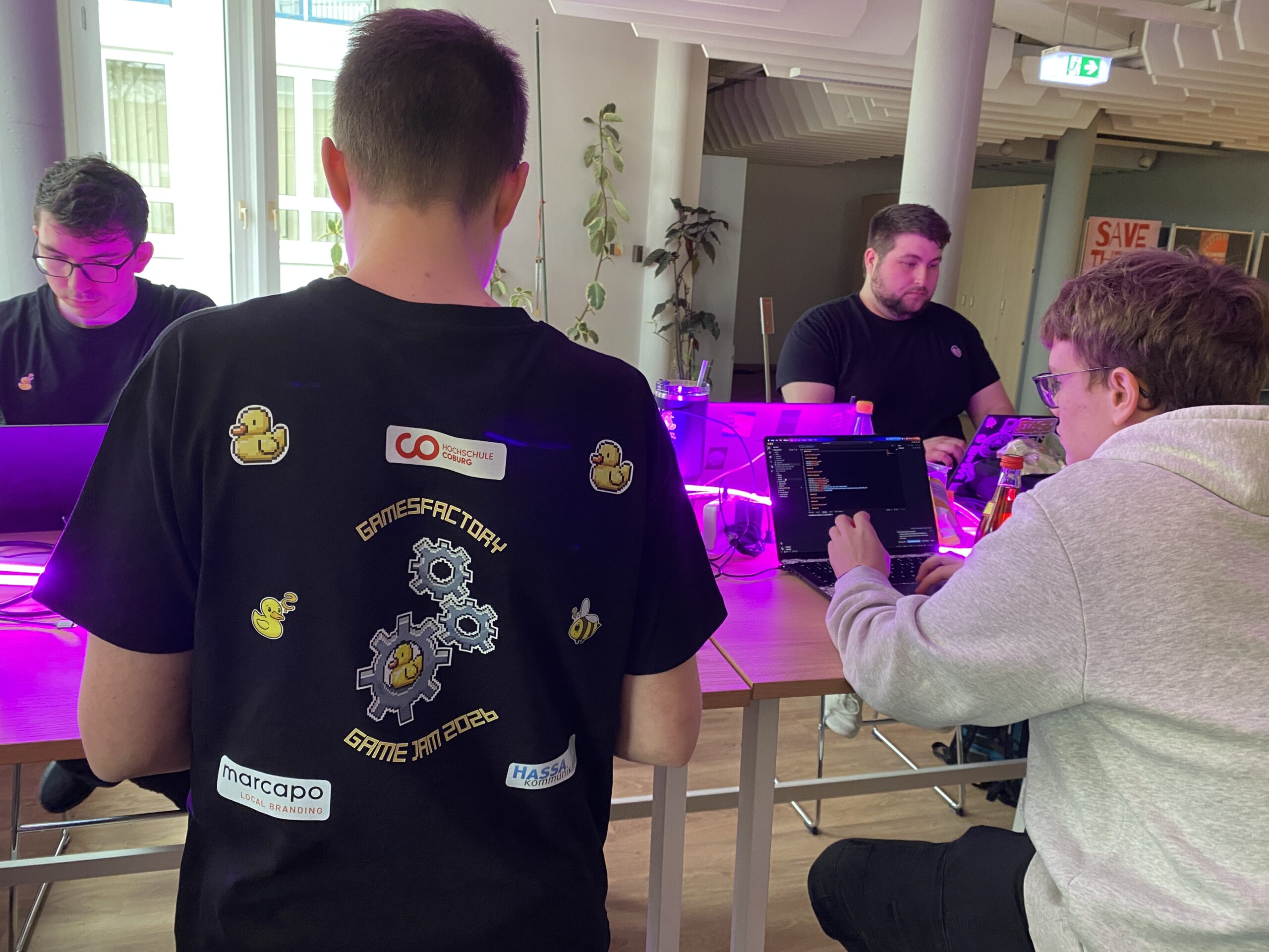 Vier Personen sitzen an einem Tisch und arbeiten an Laptops. Im Vordergrund trägt eine Person ein schwarzes "Gamesfactory Game Jam 2023"-Shirt, das mit Ausrüstung und Gummienten-Aufklebern verziert ist und den Geist von "Fail to Success" im Vorfeld des Game Jam 2026 verkörpert. at Hochschule Coburg