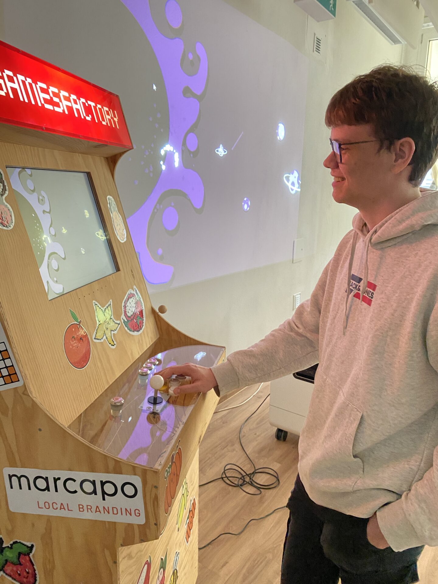 Ein junger Mann mit Brille und grauem Kapuzenpulli spielt auf dem Game Jam 2026 ein Retro-Arcade-Spiel an einem verzierten Holzschrank. Auf dem Bildschirm ist ein Weltraum-Shooter-Spiel zu sehen, bunte Aufkleber bedecken das Gehäuse, und an der Wand daneben leuchtet ein Projektor. at Hochschule Coburg