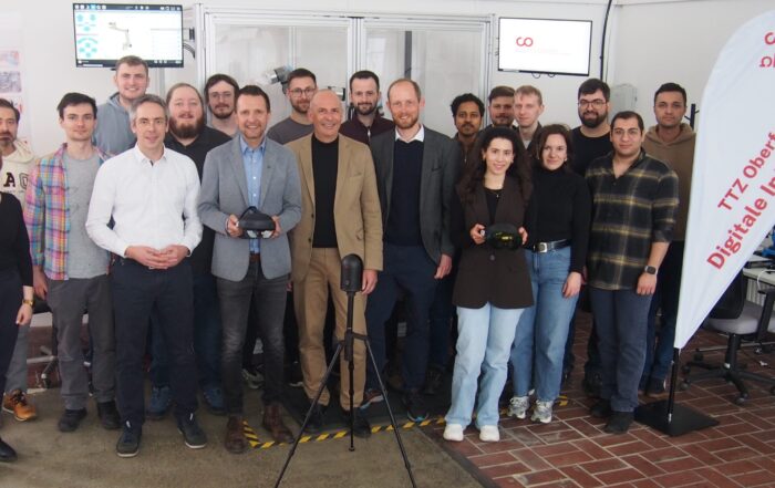 Eine Gruppe von etwa 20 Personen, Männer und Frauen, steht in einem Innenraum zusammen und lächelt in die Kamera. Einige halten technische Geräte in der Hand, und auf einem Transparent steht "Digitale Intelligenz" als Hinweis auf die industrielle Zukunft der Region. Im Hintergrund sind Monitore und Geräte zu sehen. at Hochschule Coburg