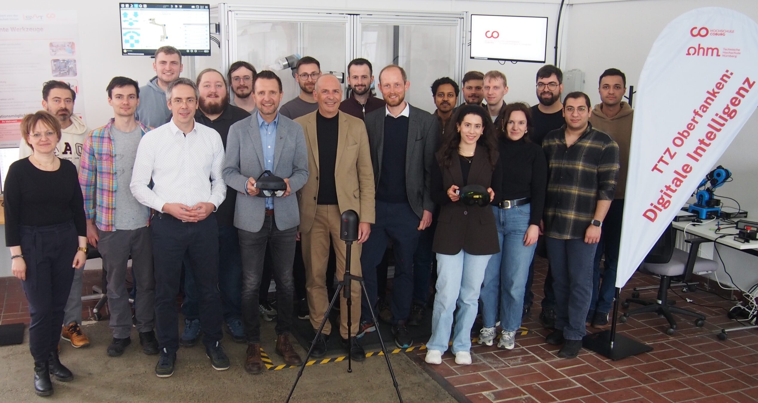 Eine Gruppe von etwa 20 Personen, Männer und Frauen, steht in einem Innenraum zusammen und lächelt in die Kamera. Einige halten technische Geräte in der Hand, und auf einem Transparent steht "Digitale Intelligenz" als Hinweis auf die industrielle Zukunft der Region. Im Hintergrund sind Monitore und Geräte zu sehen. at Hochschule Coburg