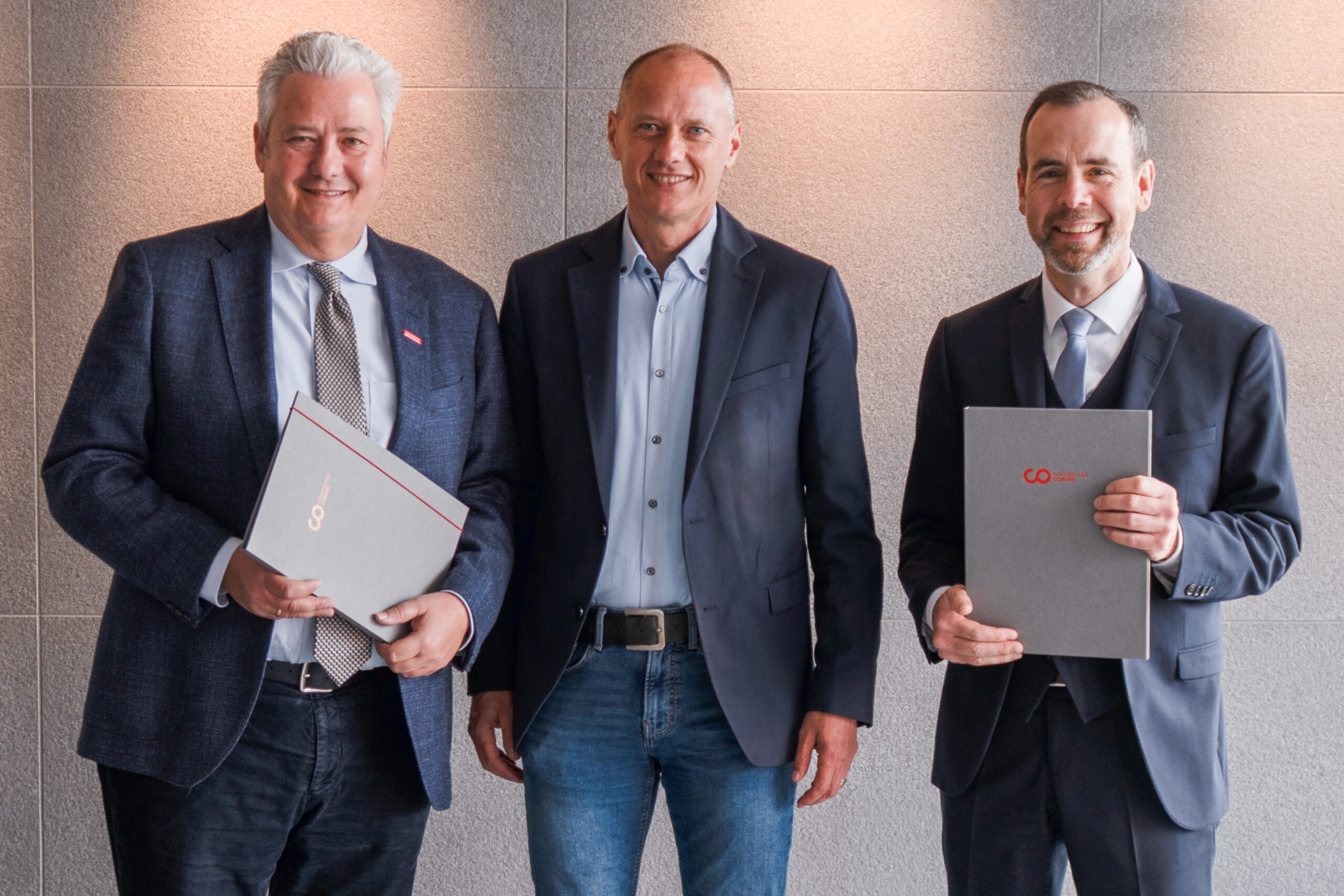 Drei Männer in Geschäftskleidung stehen lächelnd vor einer grau strukturierten Wand, möglicherweise bei einer Veranstaltung von Uropas Wissen zum Thema aktuelle Forschung. Zwei von ihnen halten graue Mappen, während der Mann in der Mitte die Hände an den Seiten hält, was den Anschein erweckt, dass es sich um den Studiengang Bauerhalt handelt. at Hochschule Coburg