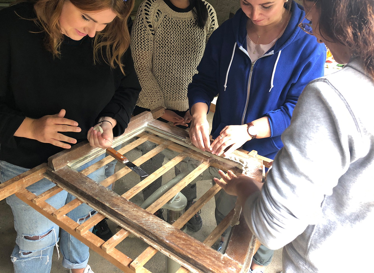 Vier Personen arbeiten gemeinsam an der Restaurierung eines hölzernen Fensterrahmens, wobei sie Uropas Wissen mit Bauerhalt-Techniken kombinieren und Werkzeuge wie Pinsel und Schleifpapier verwenden. Der Fokus liegt auf ihren Händen und dem Projekt, nicht auf ihren Gesichtern. at Hochschule Coburg