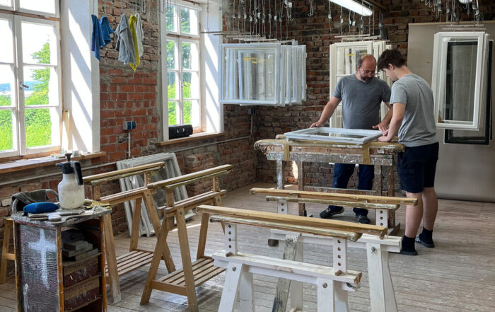 Zwei Personen stehen an einem Arbeitstisch in einer hellen Werkstatt mit großen Fenstern, freiliegenden Ziegelwänden und Holzbearbeitungswerkzeugen um sie herum und arbeiten gemeinsam an einem Projekt, das von der aktuellen Forschung und von Bauerhalt-Techniken inspiriert ist. at Hochschule Coburg