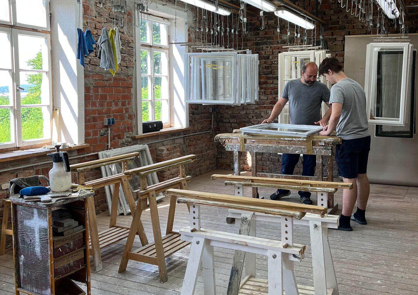 Zwei Personen stehen an einem Arbeitstisch in einer hellen Werkstatt mit großen Fenstern, freiliegenden Ziegelwänden und Holzbearbeitungswerkzeugen um sie herum und arbeiten gemeinsam an einem Projekt, das von der aktuellen Forschung und von Bauerhalt-Techniken inspiriert ist. at Hochschule Coburg