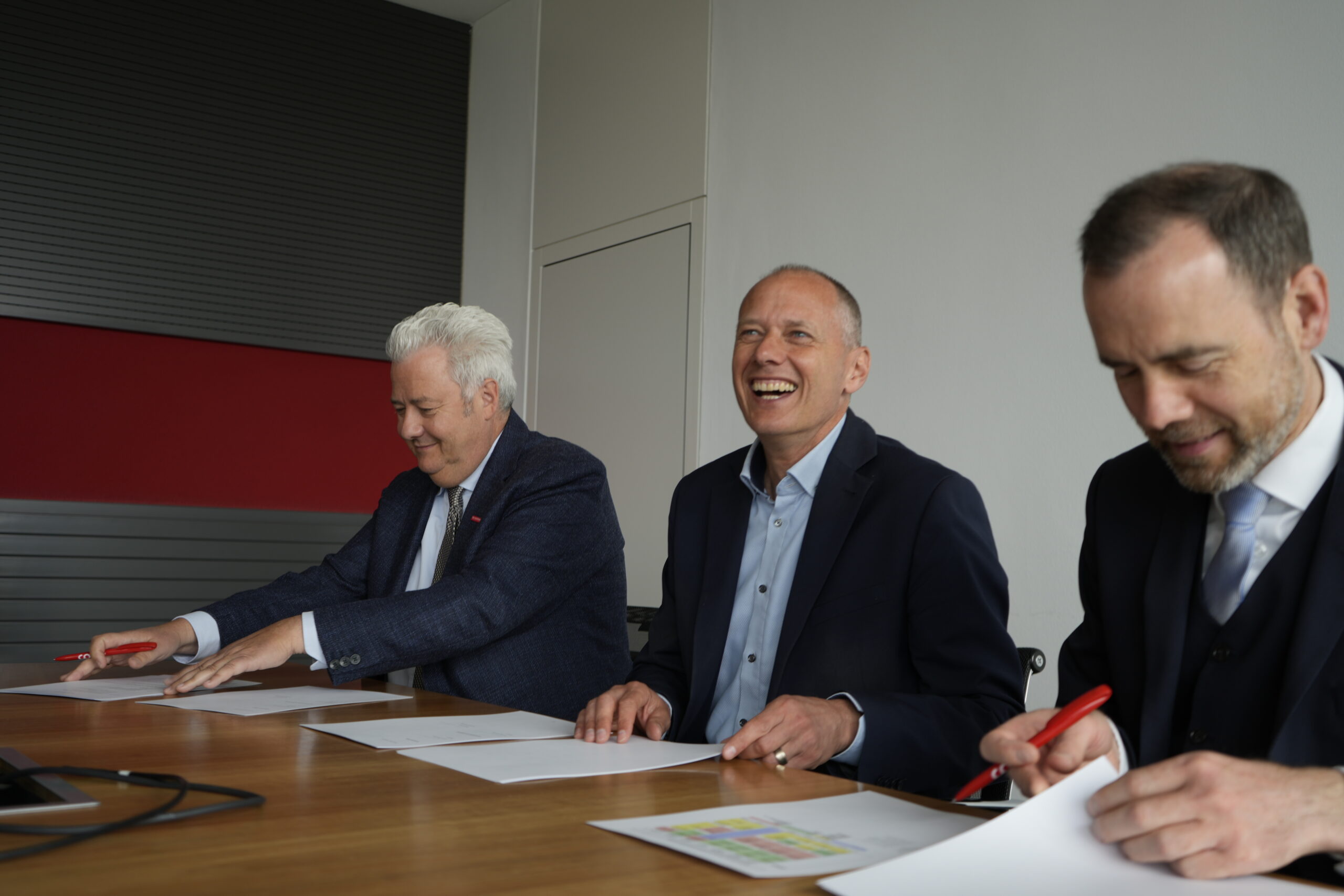 Drei Männer in Anzügen sitzen an einem Tisch, lächeln und unterschreiben Dokumente mit roten Stiften. Die Stimmung wirkt freundlich und kollegial im modernen Bauerhalt-Büro, wo aktuelle Forschung auf zeitlose Geschäftstradition trifft. at Hochschule Coburg