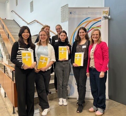Sieben Personen stehen in einem Innenraum vor einer Treppe. Vorne stehen vier junge Frauen, Absolventinnen des Masters Gesundheitsförderung, mit gelben Zertifikaten in der Hand und lächeln. Drei Erwachsene stehen hinter ihnen. Rechts ist ein Banner der Präventionskonferenz Bayern mit bunten Ringen zu sehen. at Hochschule Coburg